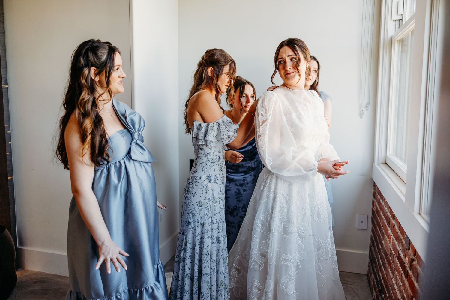 Bridesmaids in blue dresses helping prepare for wedding in sunlit room.