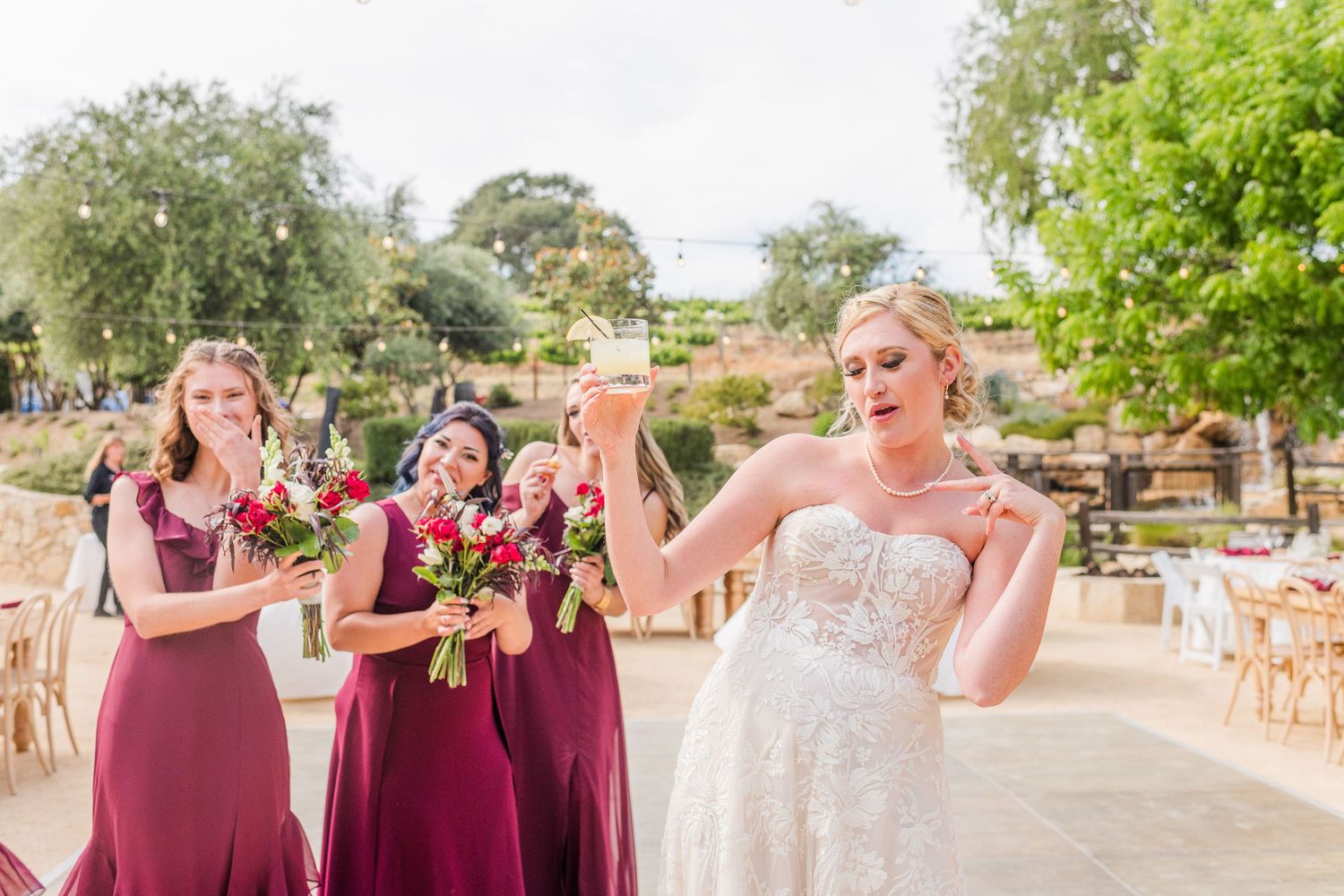 Bride celebrating with bridesmaids in burgundy dresses at outdoor venue.