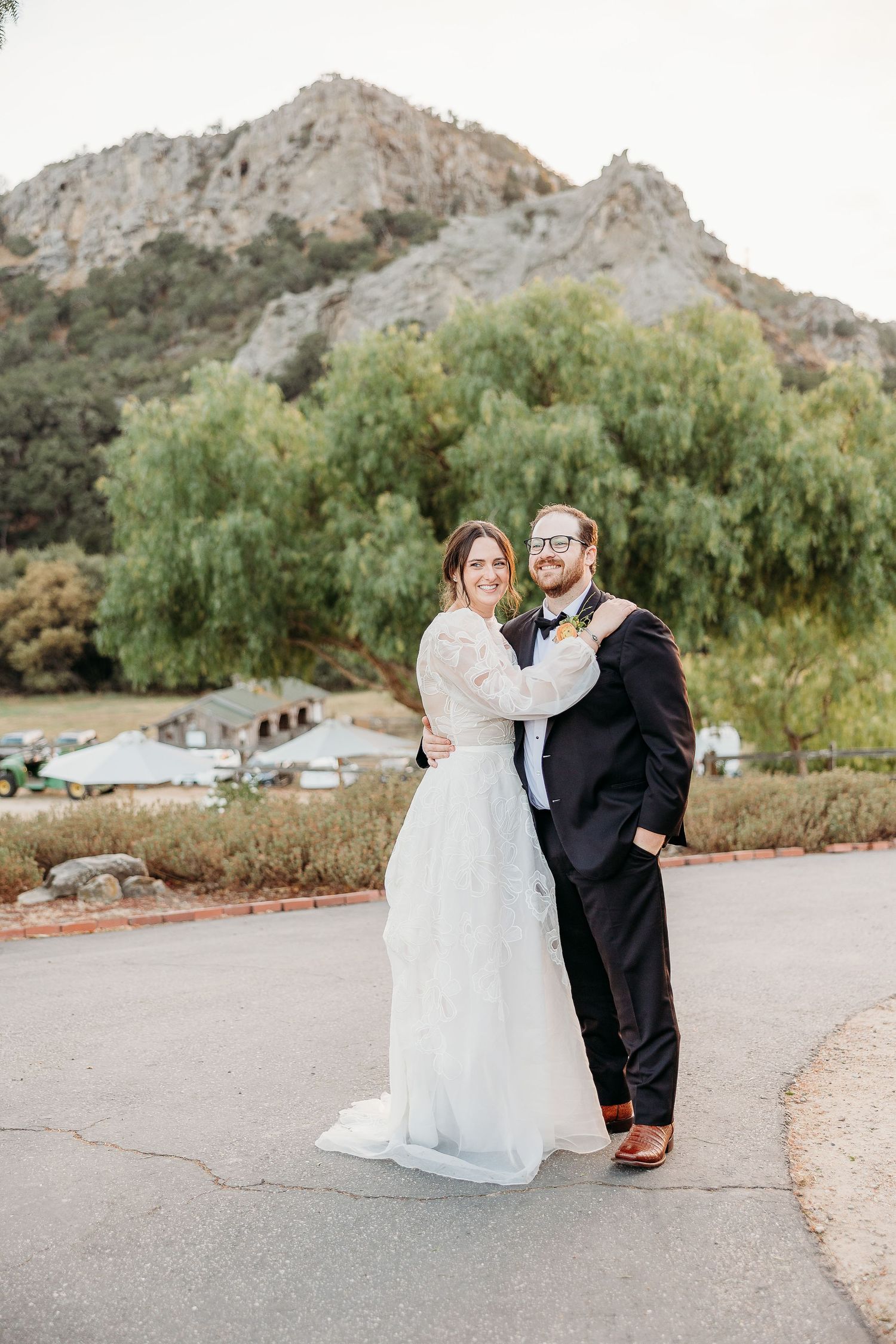 A couple shares a romantic moment in their wedding attire against a mountainous backdrop with willow trees.