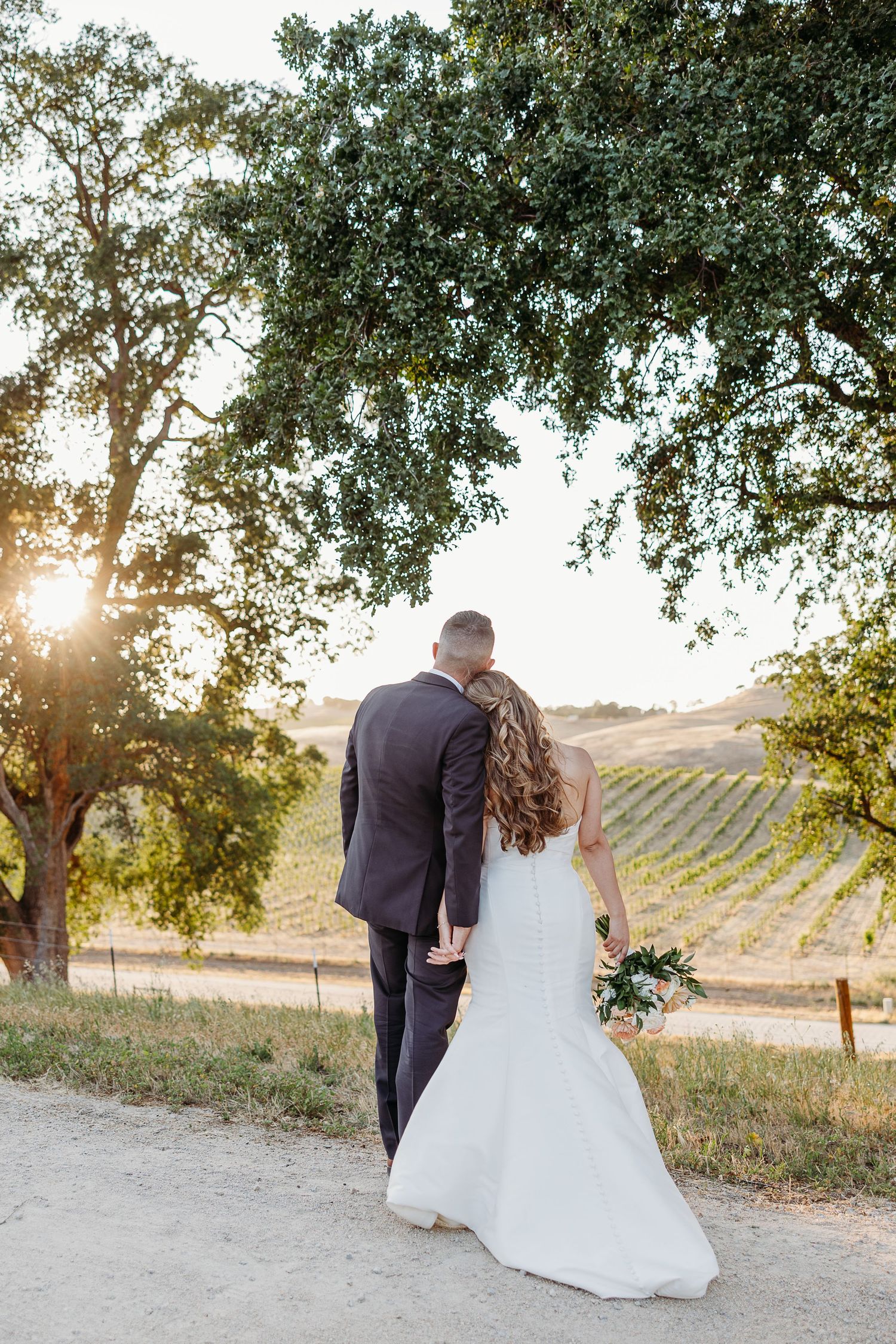 Elegant newlyweds walk through a vineyard at golden hour under sprawling oak trees.