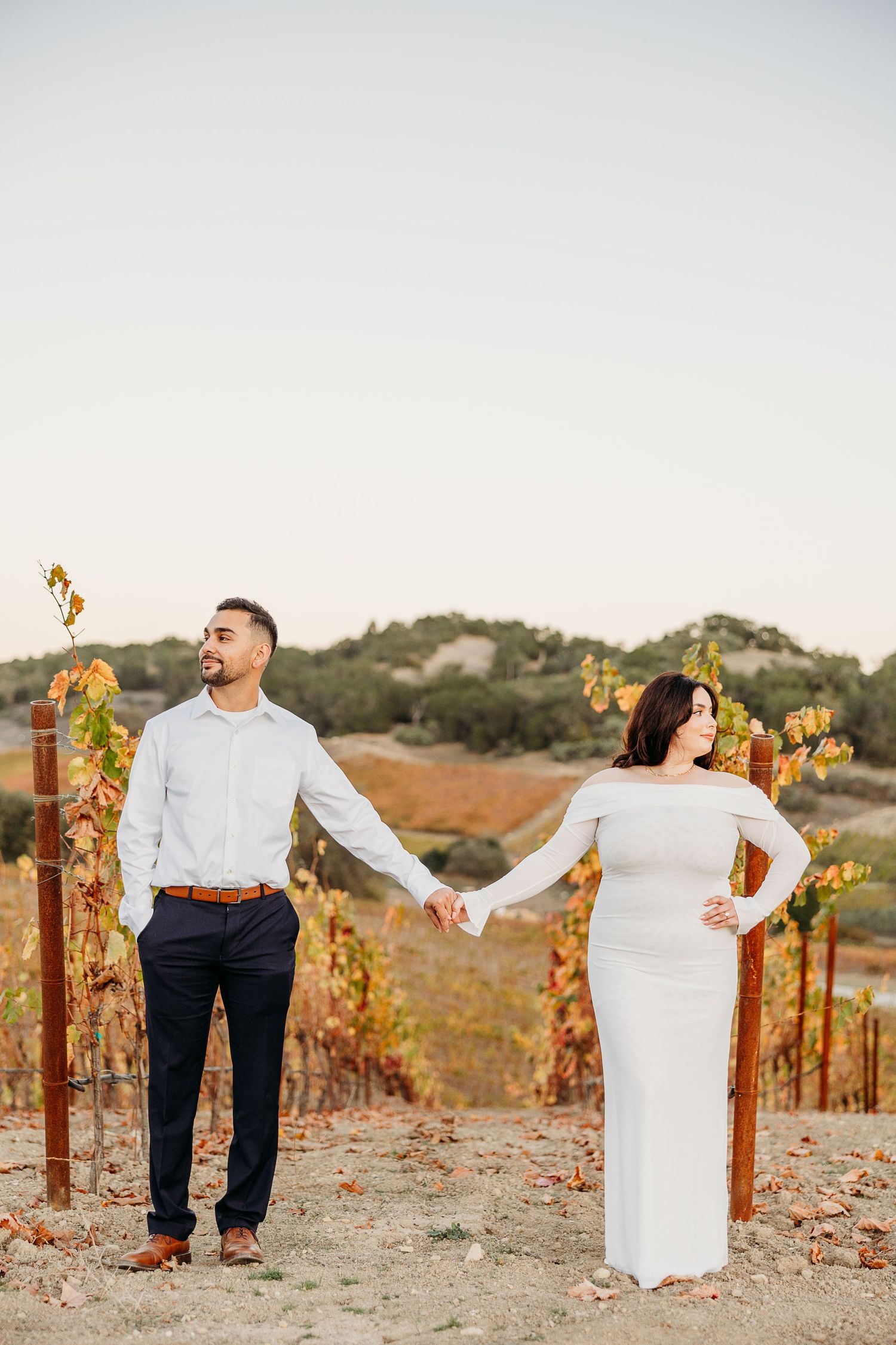 A couple holds hands while standing among autumn vineyard rows at sunset.