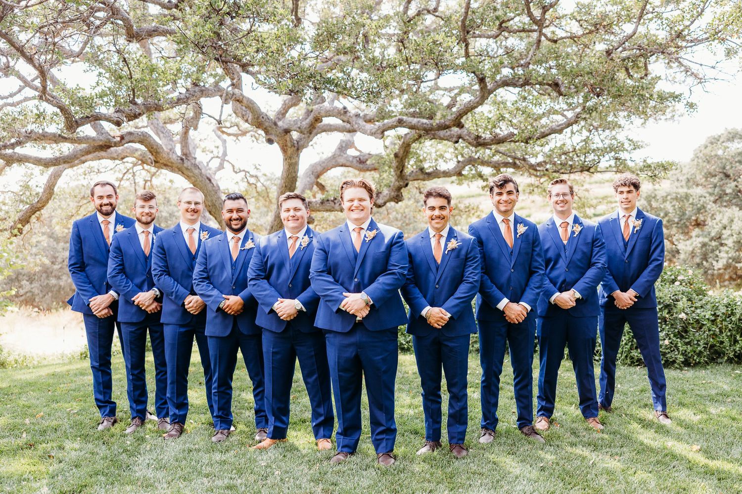 Groomsmen lined up in matching navy blue suits against a scenic outdoor backdrop.