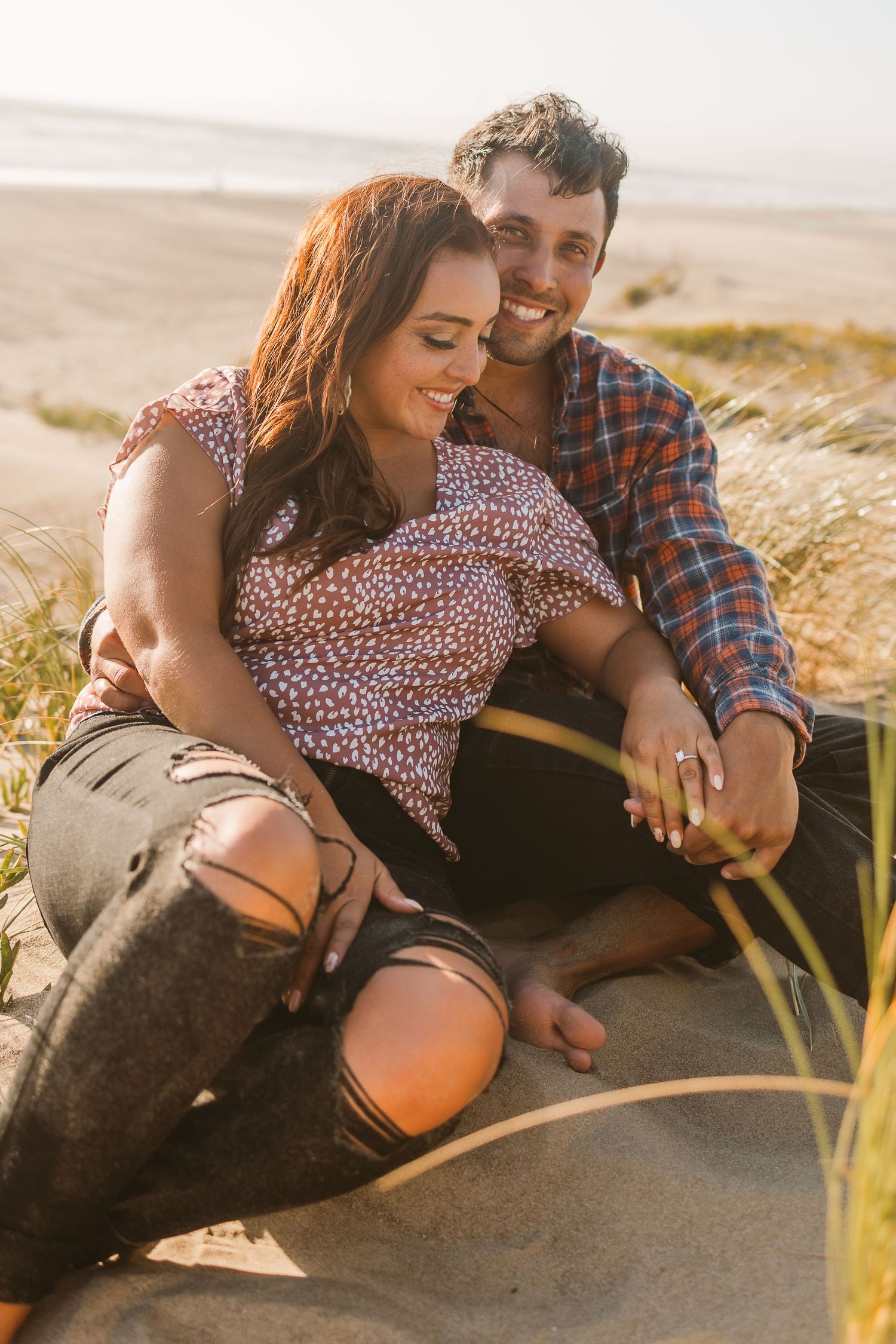 Couple sitting together on sandy beach with beach grass in foreground.