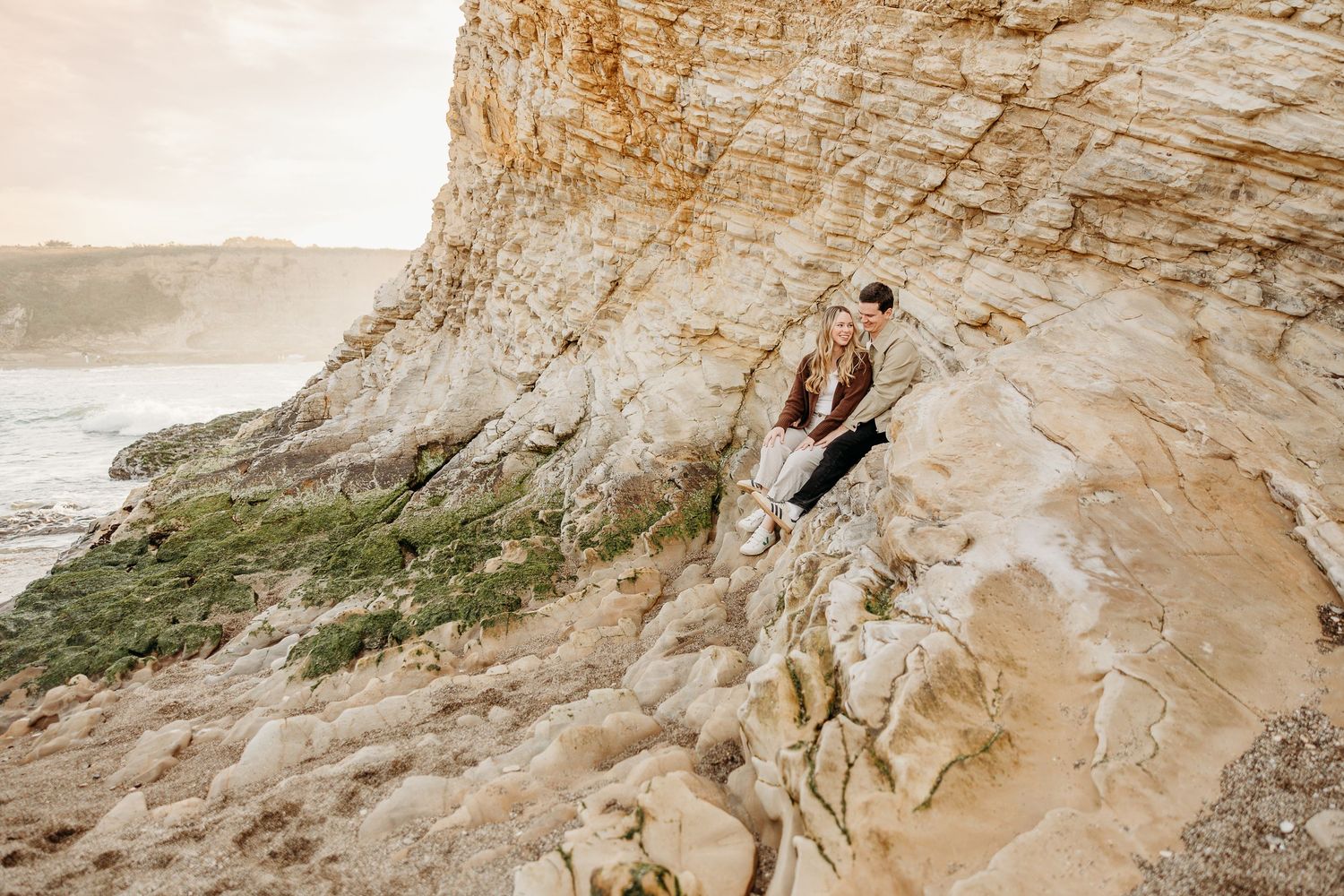 Two people explore rocky coastal cliffs during a sunset photoshoot.