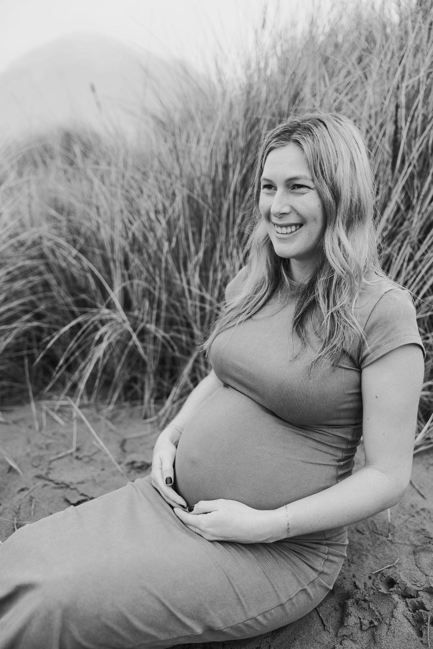 Black and white maternity photo captures an expectant mother sitting amid beach grass with a joyful expression.