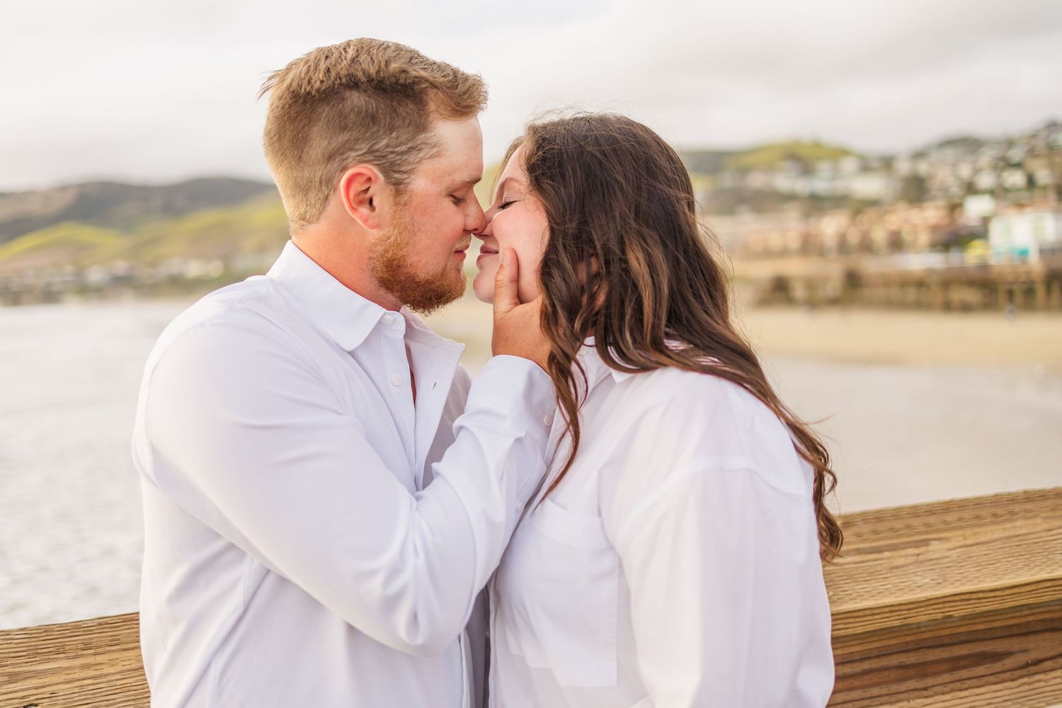 A romantic couple sharing an intimate kiss on a wooden pier overlooking coastal hills at sunset.