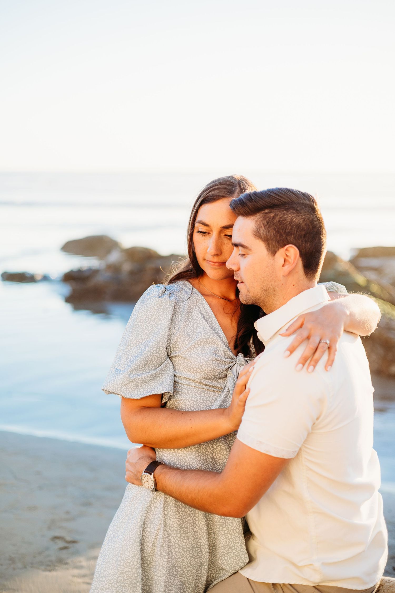 A couple embraces while sitting on coastal rocks with ocean in background.