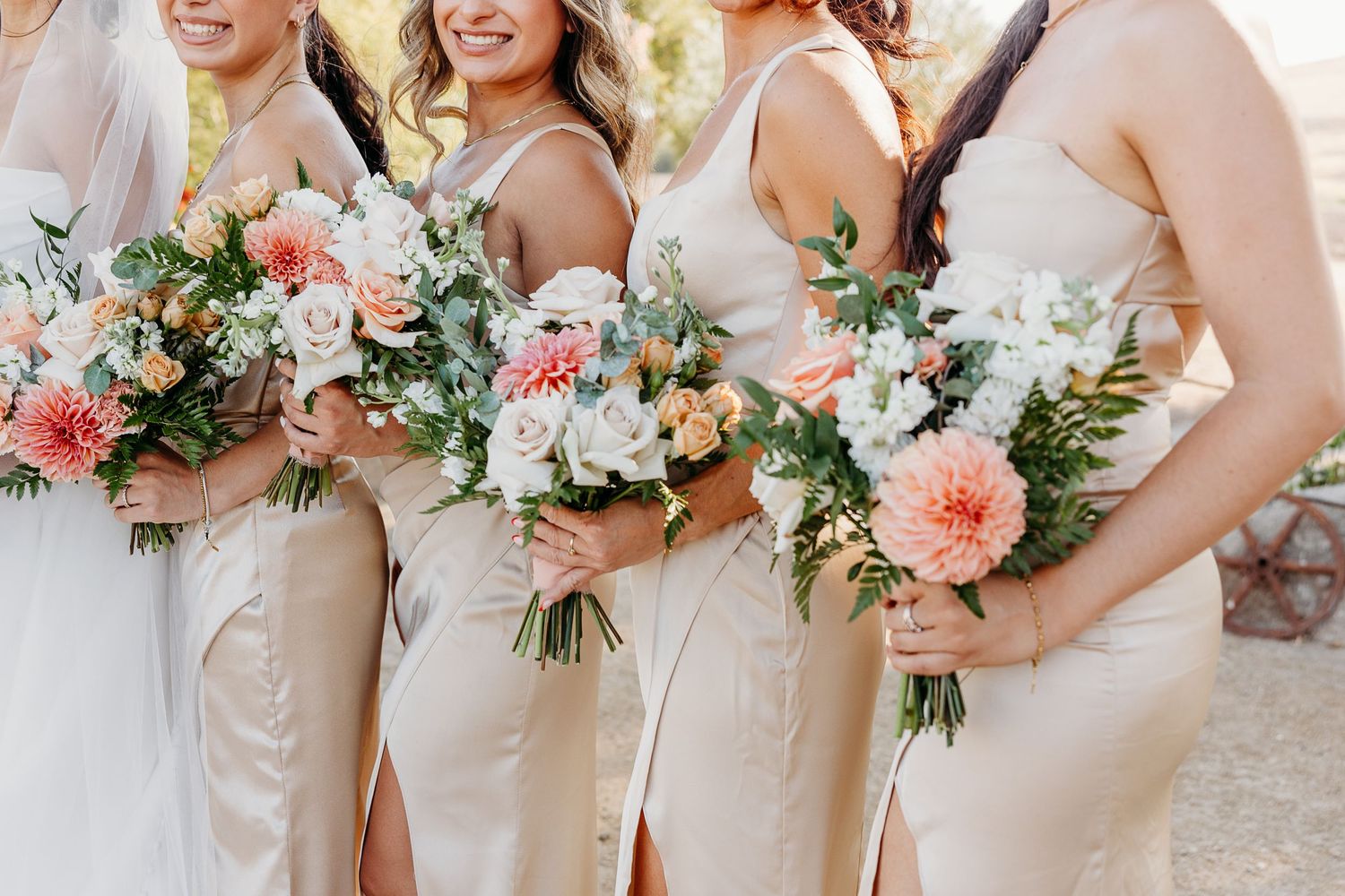 A row of bridesmaids in champagne-colored dresses hold beautiful pink and white floral bouquets at an outdoor wedding.