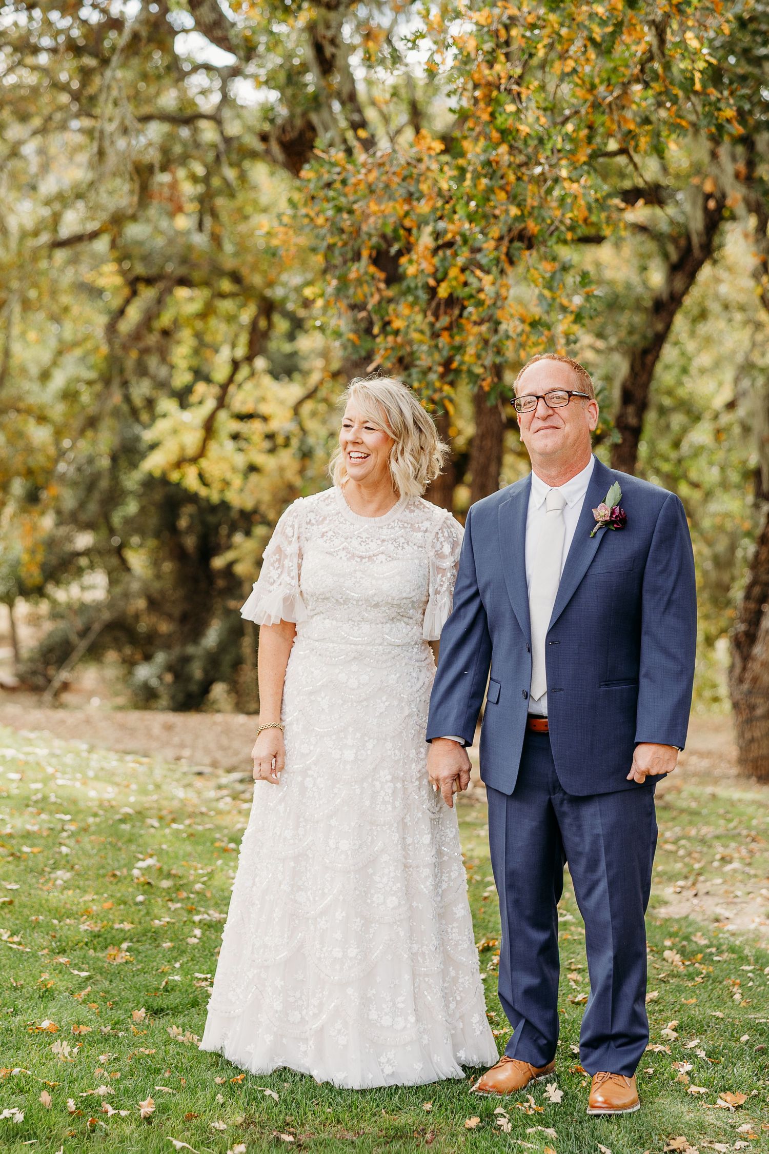 Couple walking together on grass with autumn trees in background during wedding photoshoot.