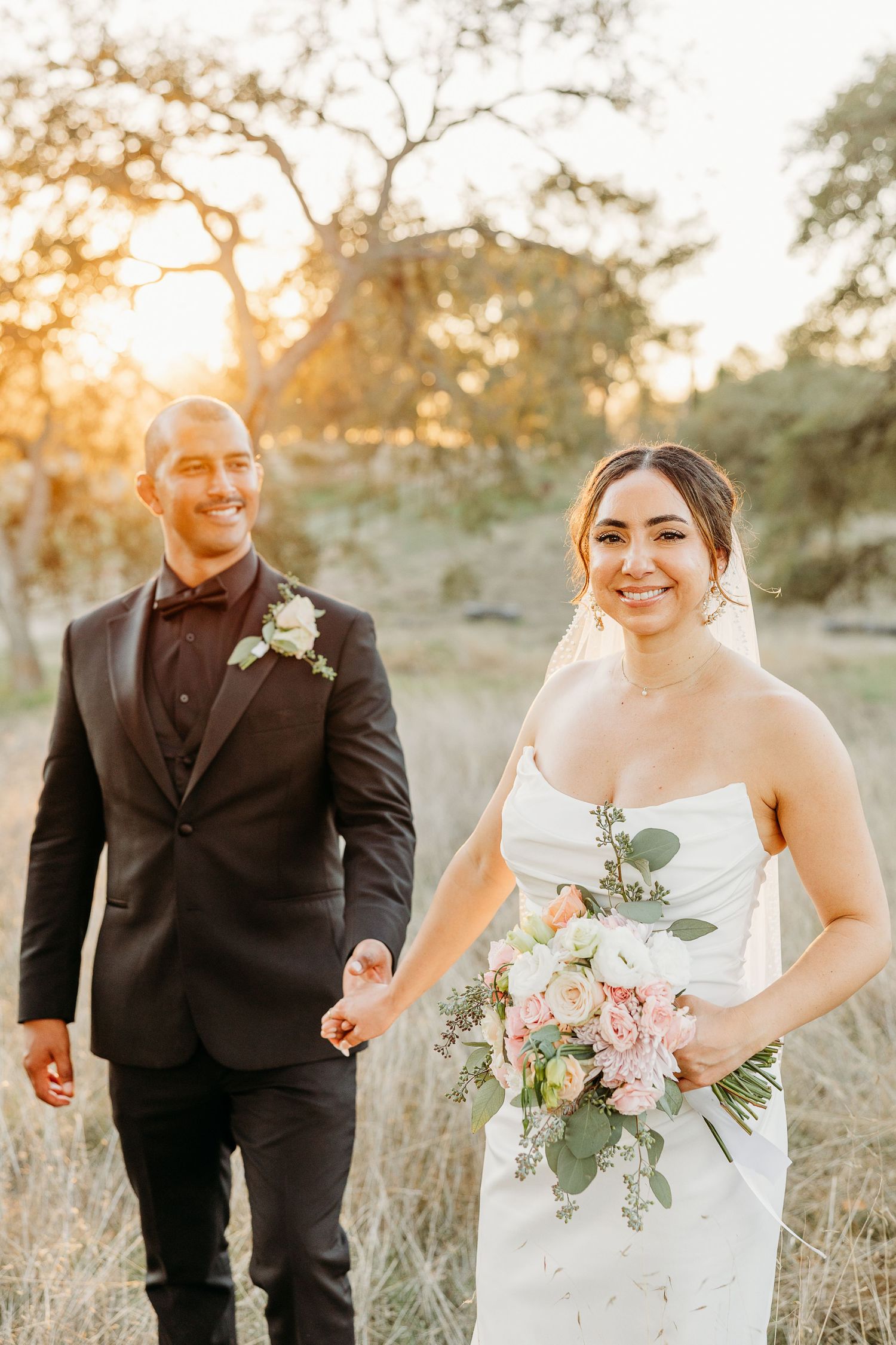Romantic wedding couple walks through golden field at sunset holding hands and carrying delicate floral bouquet.