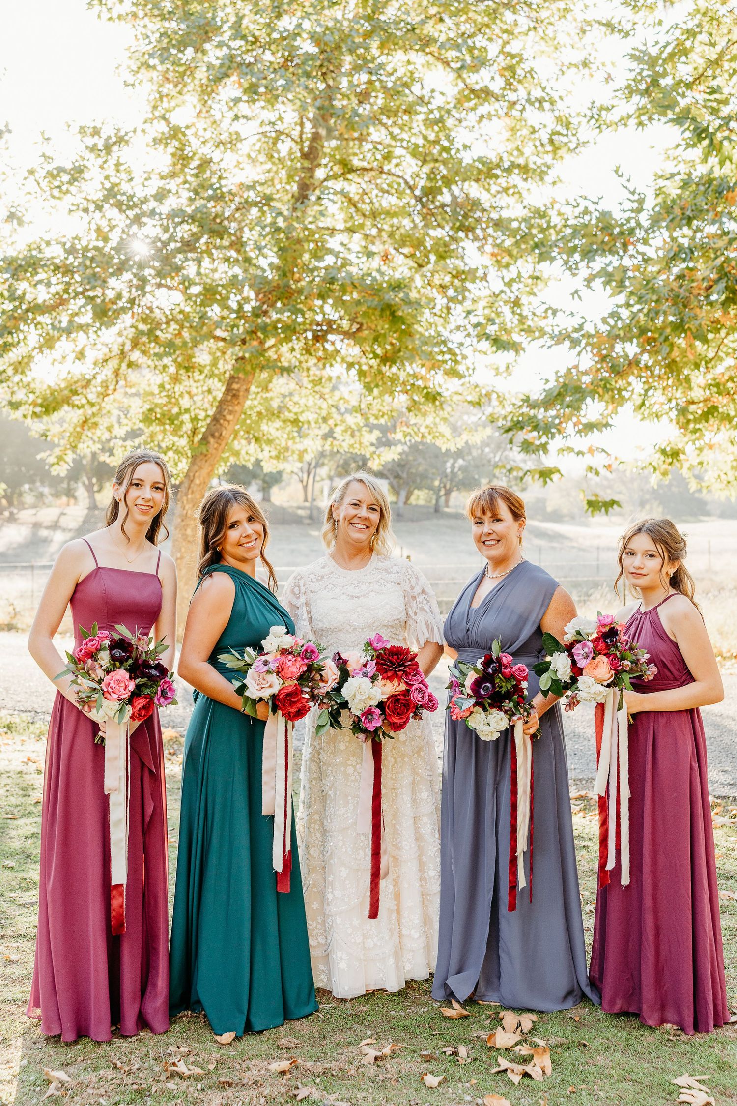 Wedding party in burgundy, teal and gray dresses pose together holding vibrant bouquets outdoors.