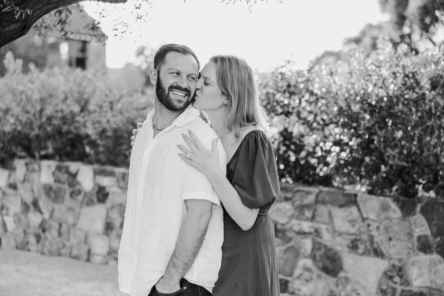 A romantic black and white photo of a couple laughing together against a stone wall backdrop.