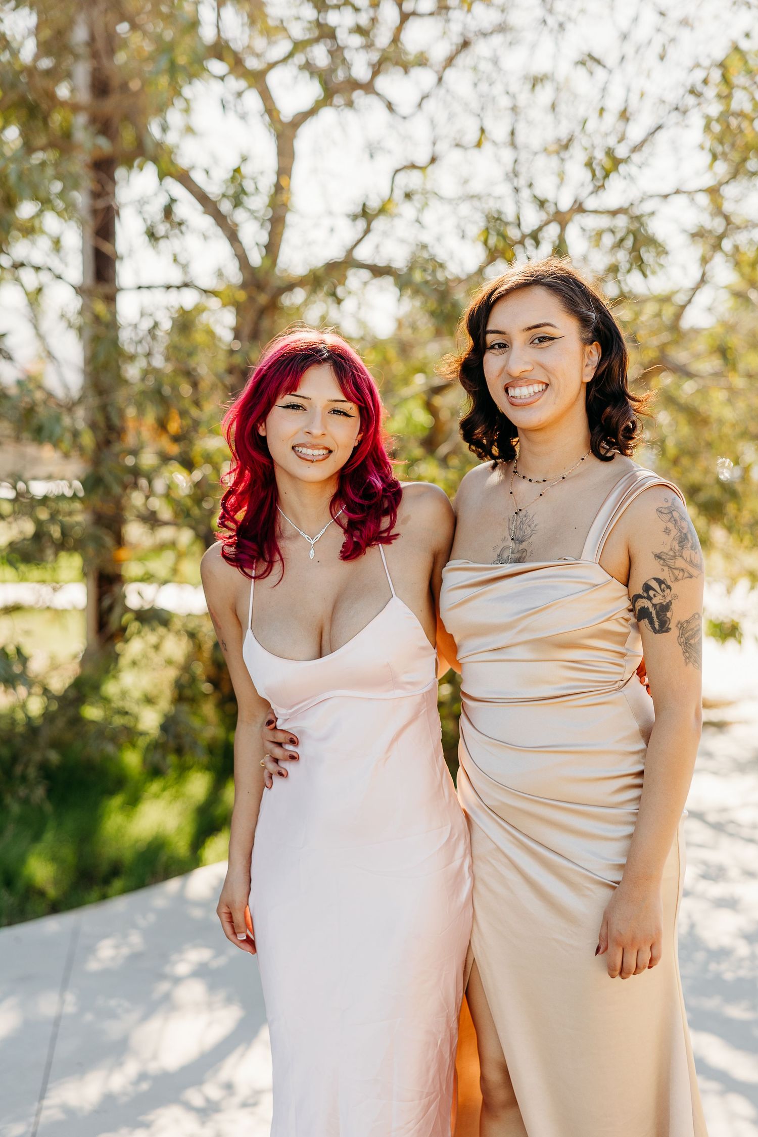 Two people in light colored dresses pose together outdoors in warm sunlight.