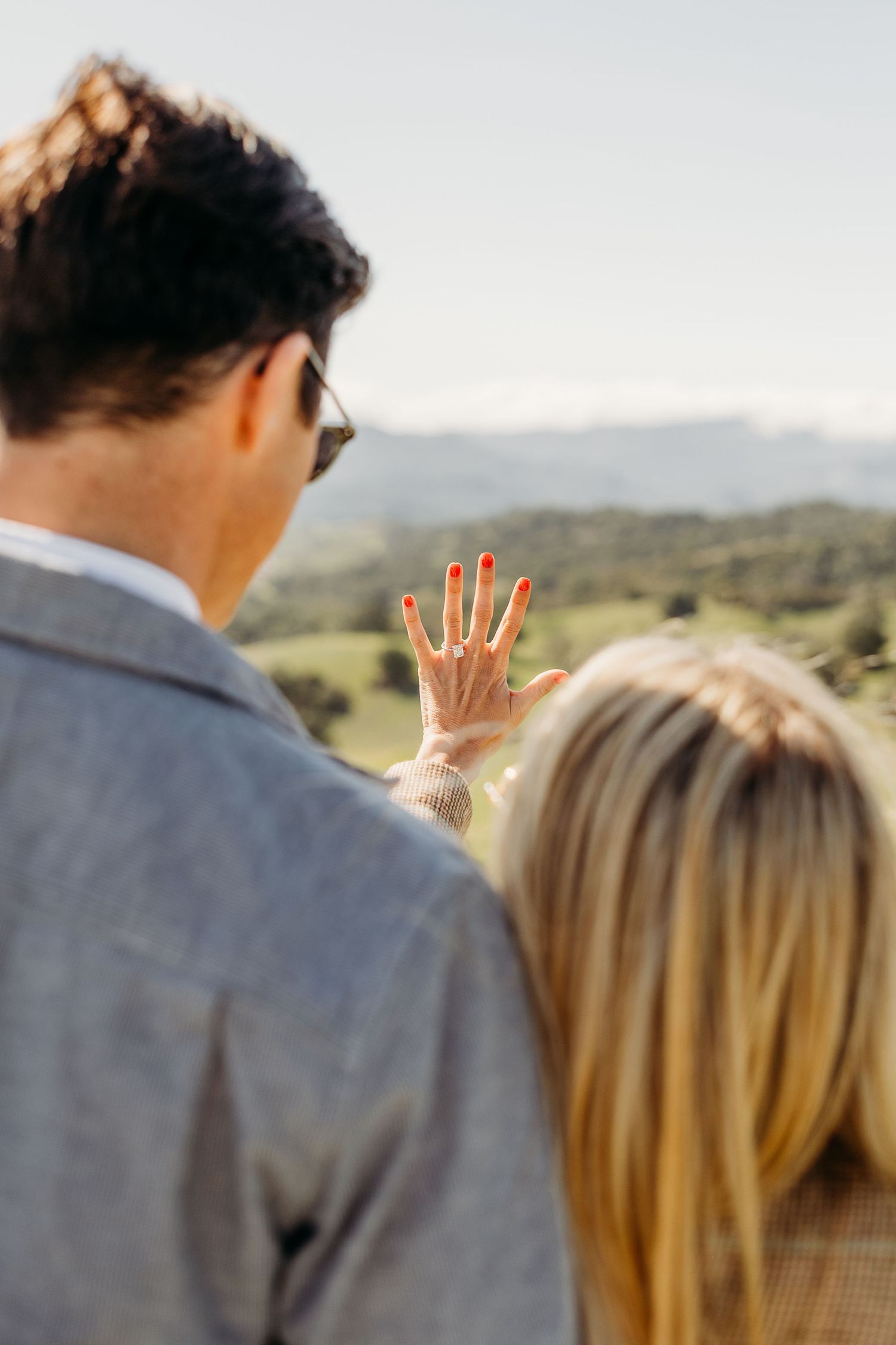 A couple in formal attire stands outdoors with their hands raised showing their fingers in a counting gesture.