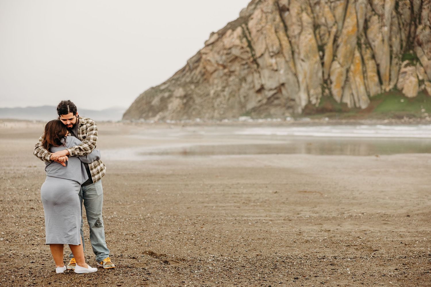 A couple embraces on a misty beach with dramatic cliffs in the background at the Pacific Northwest coast.