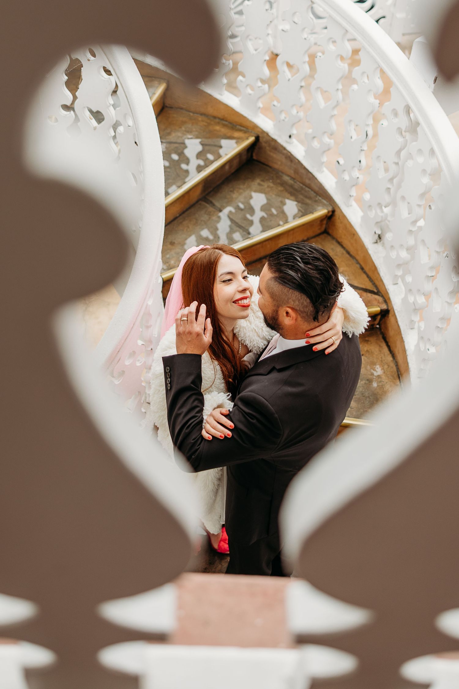 Romantic couple embraces on grand staircase with crystal chandelier and warm decor visible through heart-shaped frame.