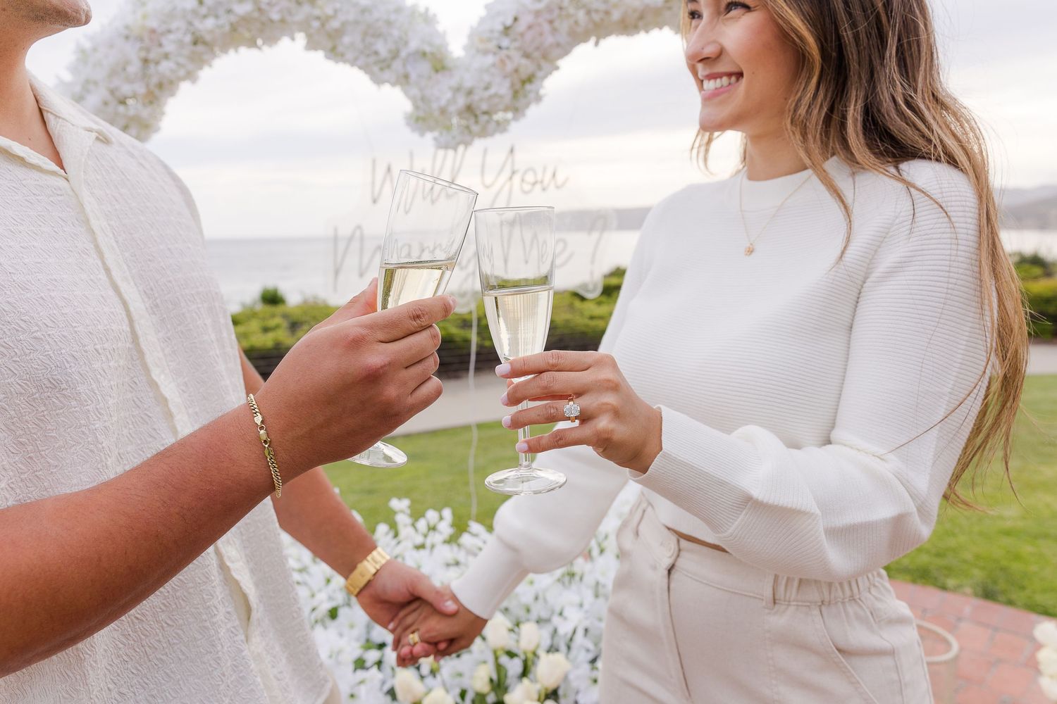 Two people toast with champagne glasses during a seaside celebration in white attire.