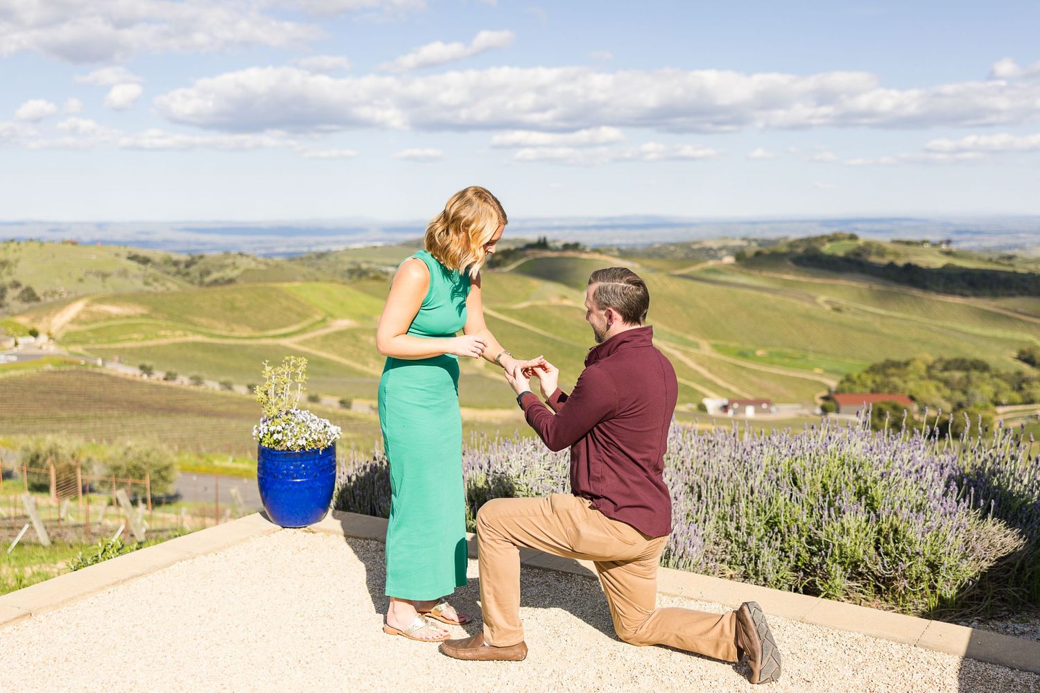 Couple sits on outdoor patio with vibrant pink neon sign and colorful flower planters against mountain backdrop.