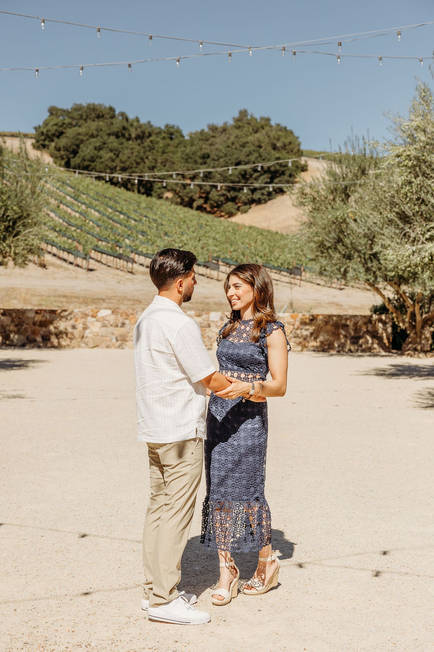 A couple dances together on a sandy path with string lights and vineyard hills in the background.