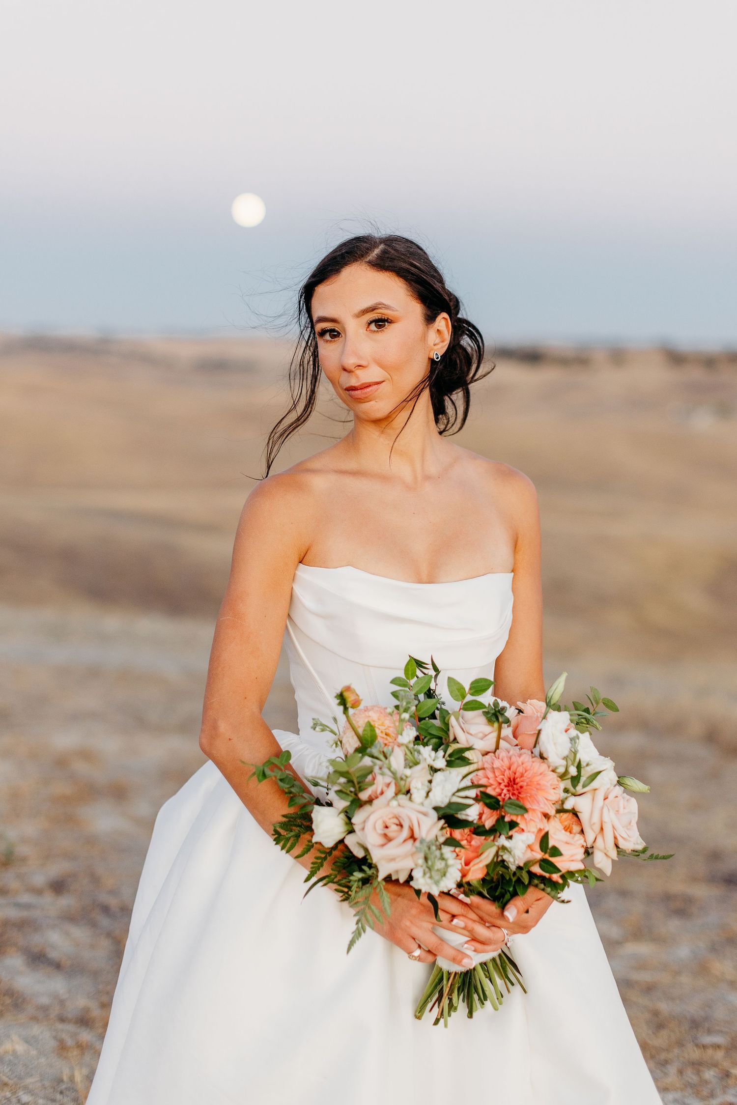 Elegant bride holds a peach and white bouquet against a desert landscape at sunset.