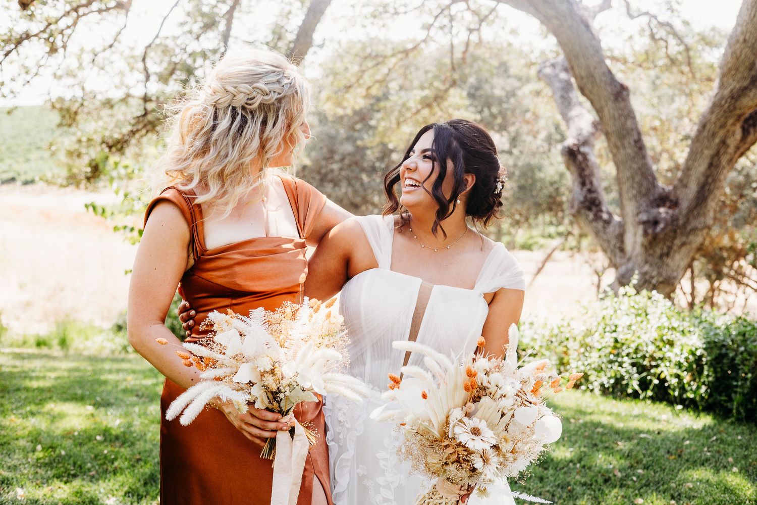 A sun-lit outdoor wedding photo with bridesmaids in rust-colored dresses holding dried flower bouquets.