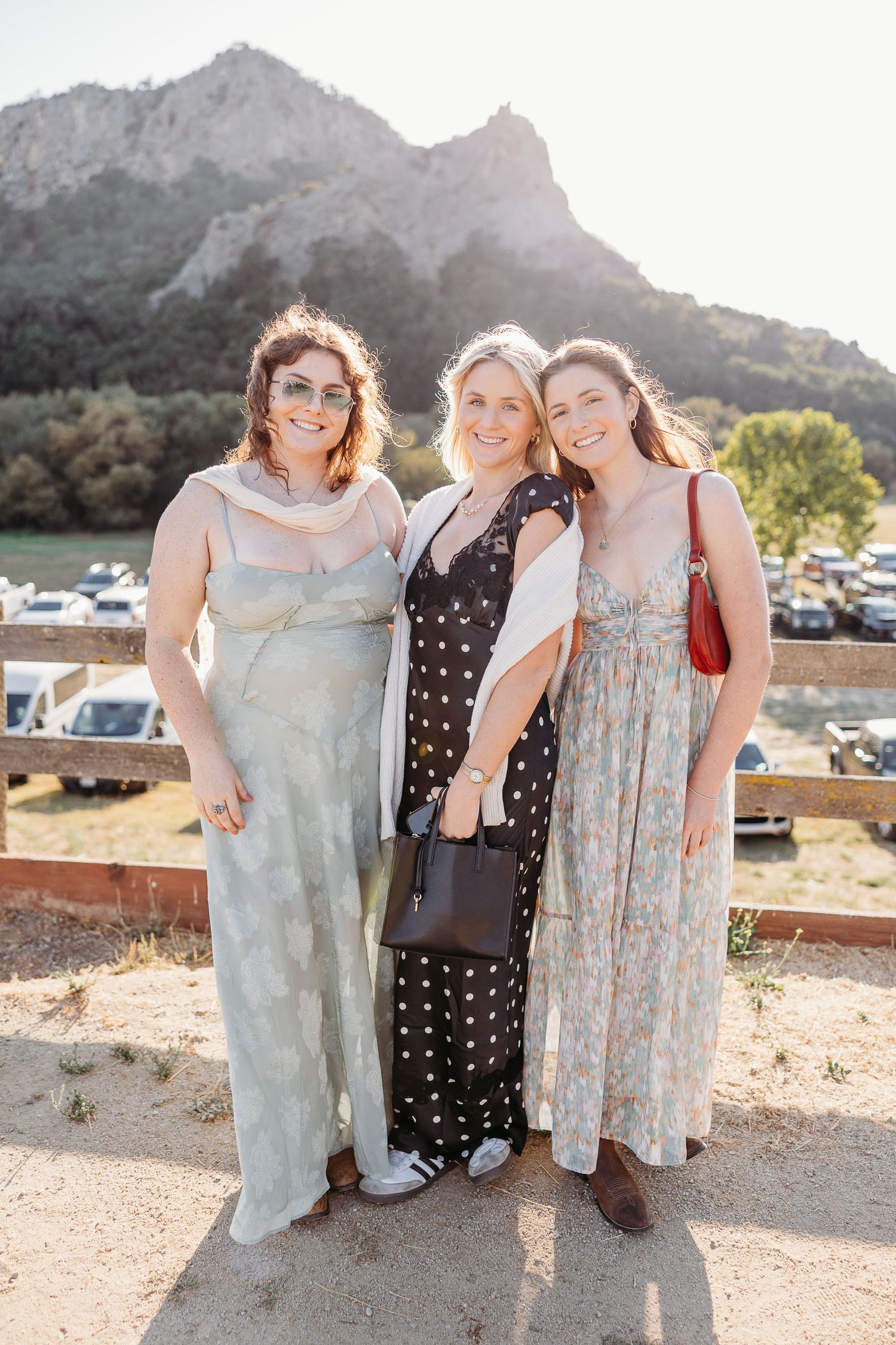 Three women in floral dresses pose together outdoors with mountains and sunlight in the background.