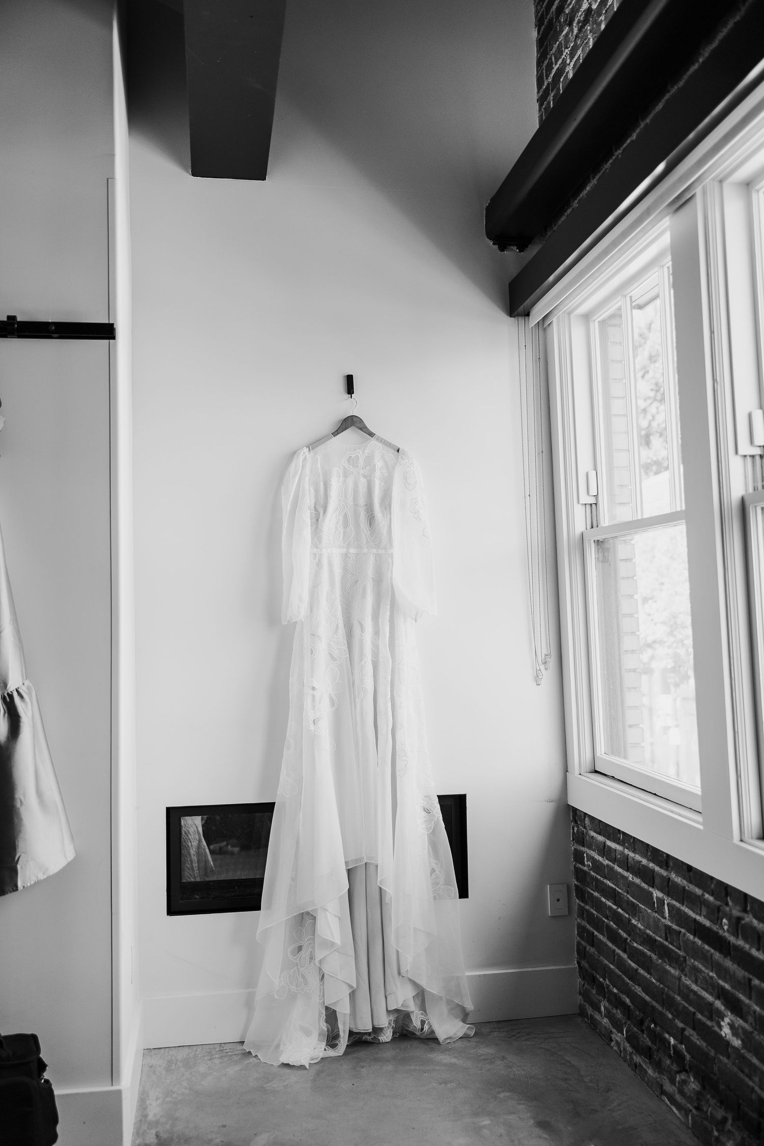 A delicate white lace wedding gown hangs elegantly against a white wall near a bright window.