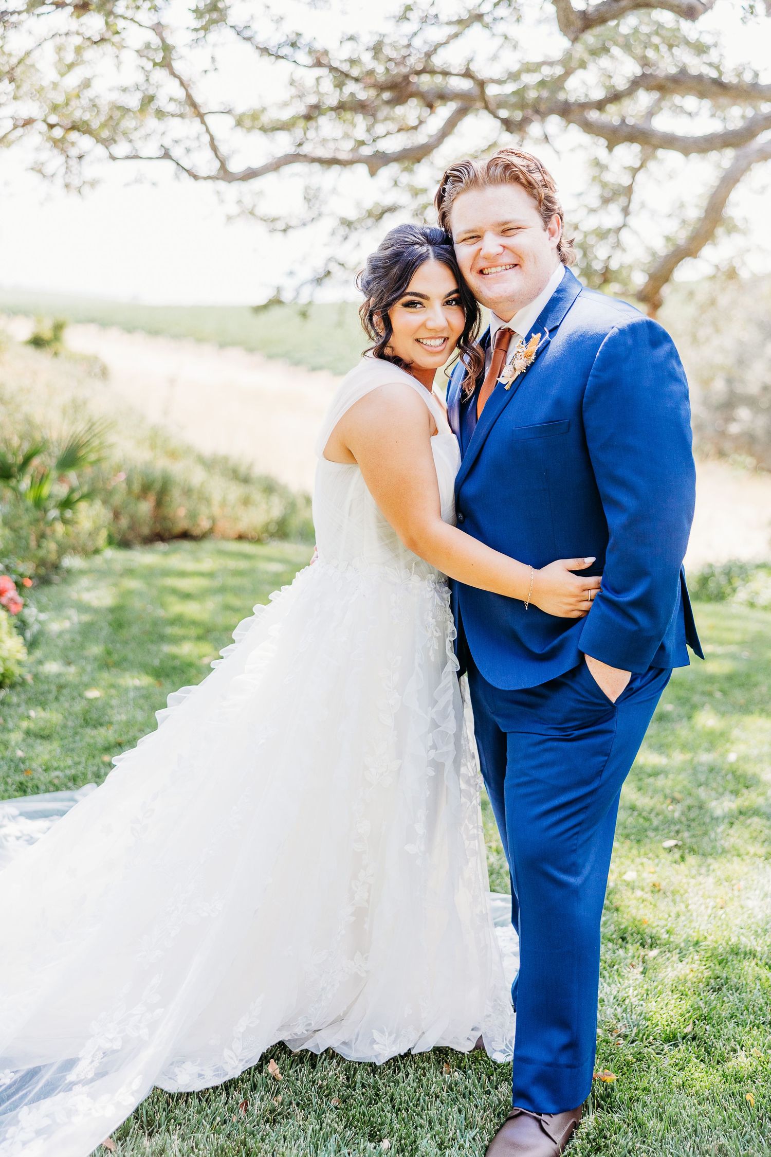 Newlyweds pose together on a hilltop, bride in white dress and groom in vibrant blue suit.