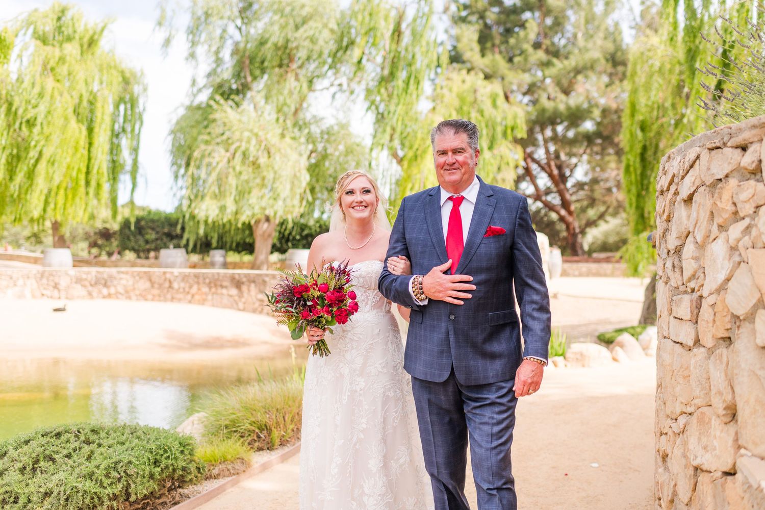 A wedding couple walks along a garden path with weeping willows and red floral bouquet.