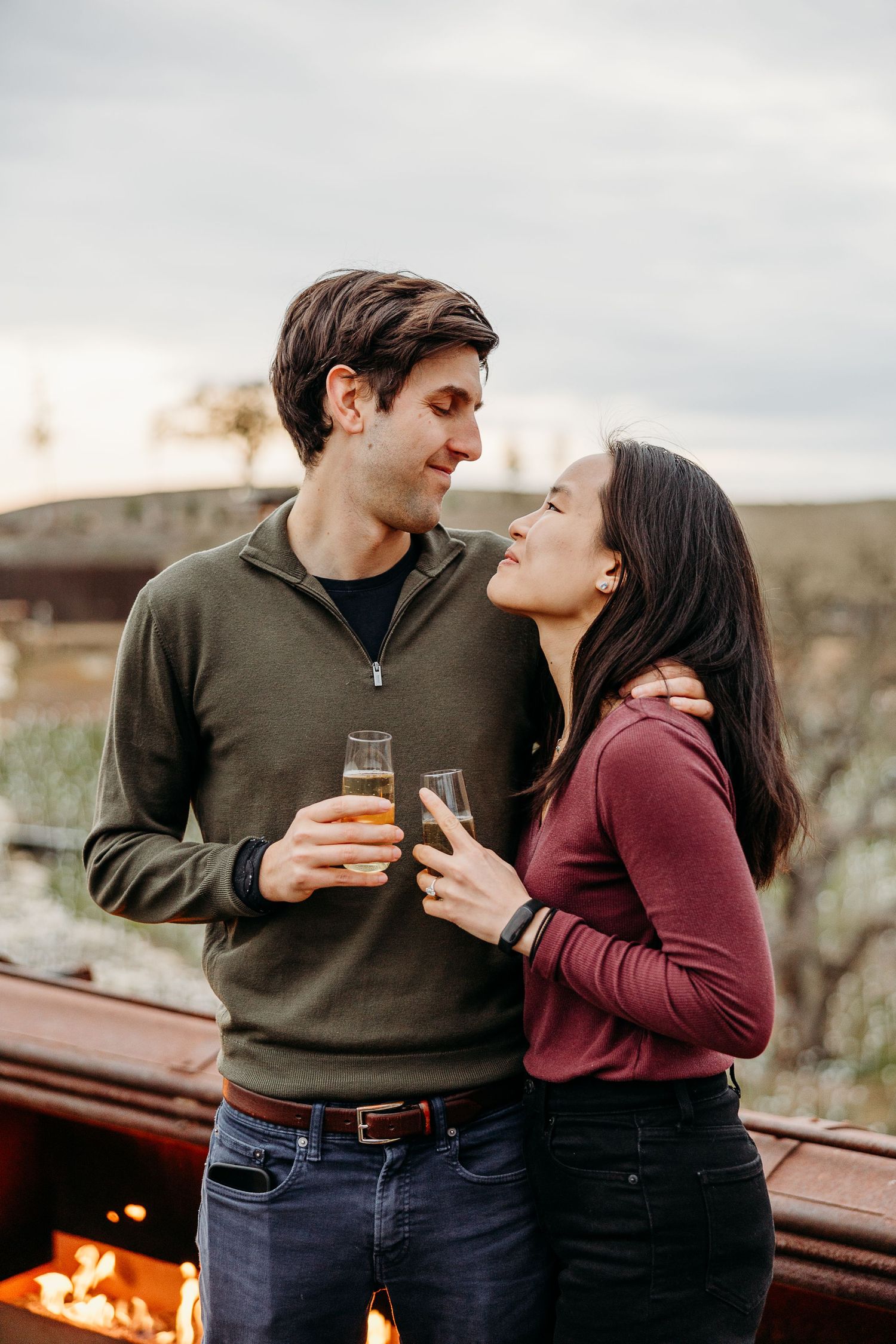 A couple enjoying drinks and laughter together by a rustic wooden railing at sunset.
