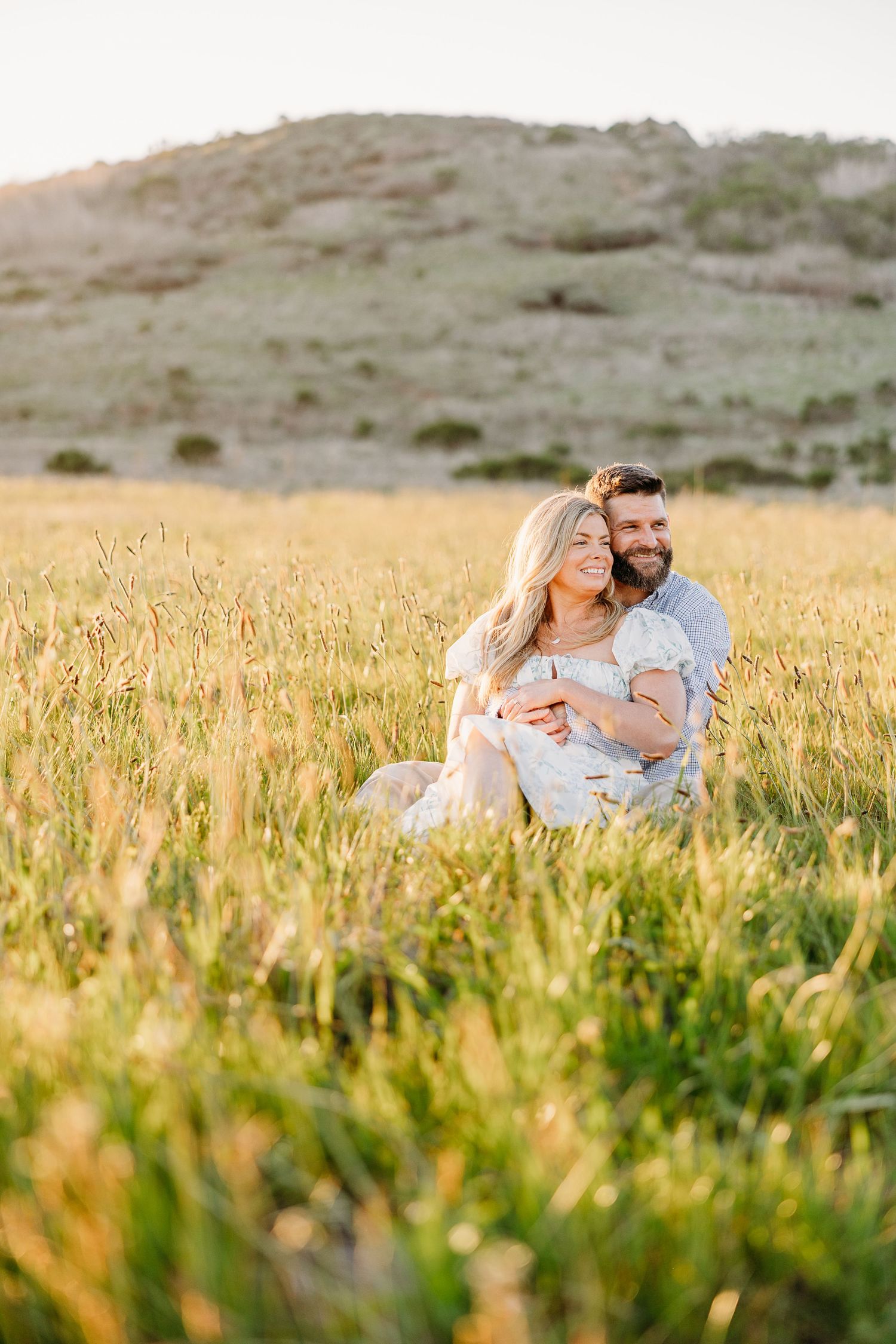 A couple embraces in a golden wheat field at sunset with rolling hills in the background.