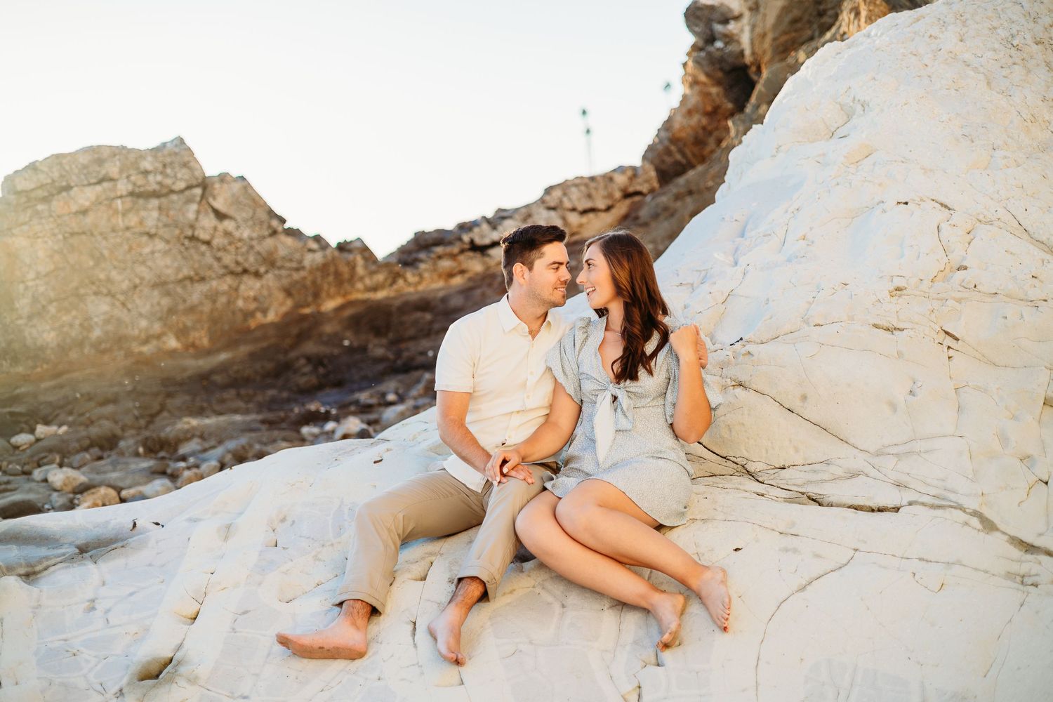 A couple laughs together while sitting on white coastal rocks at sunset.