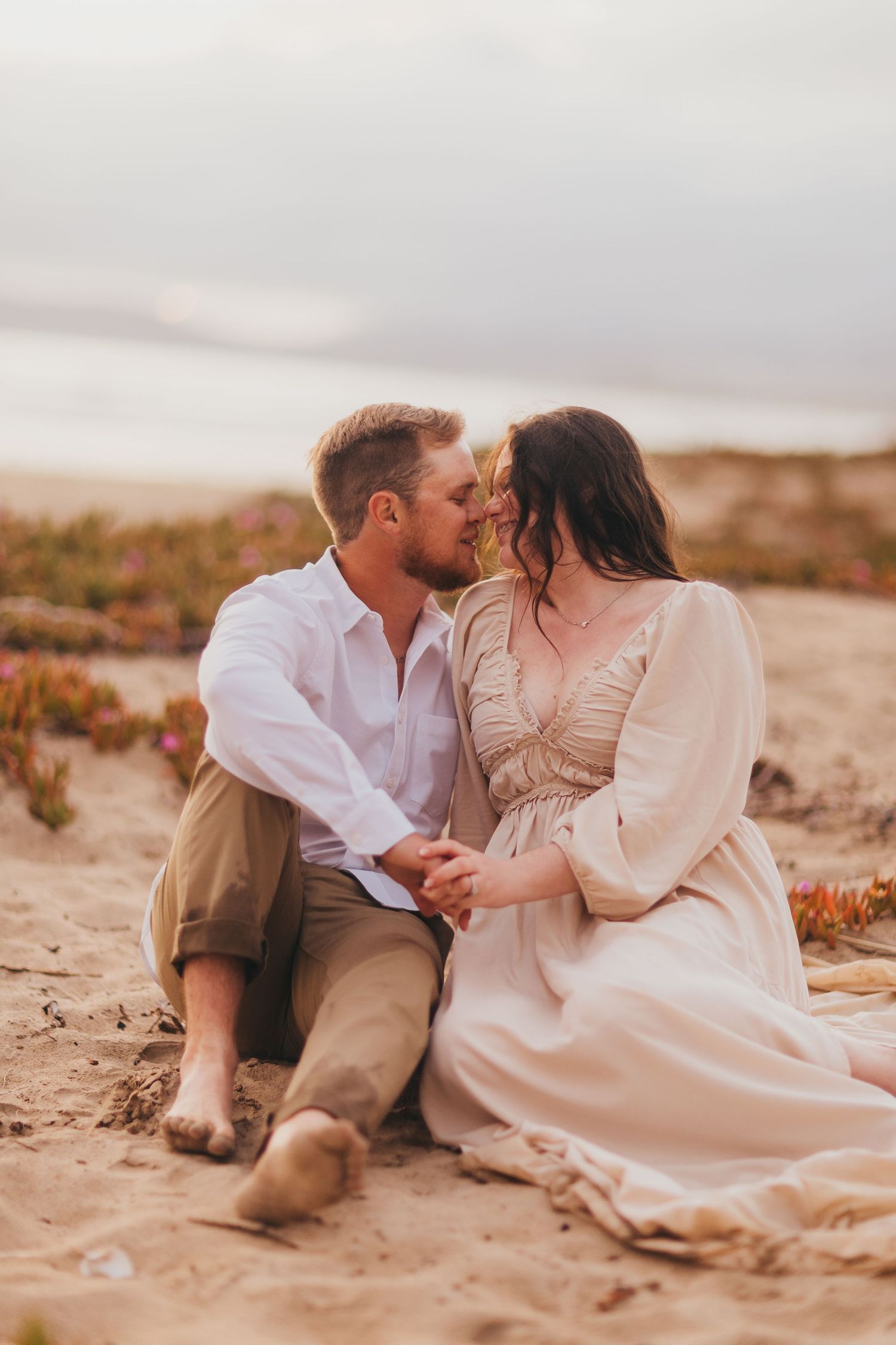 Intimate beach photoshoot featuring couple in white and beige outfits against coastal backdrop.