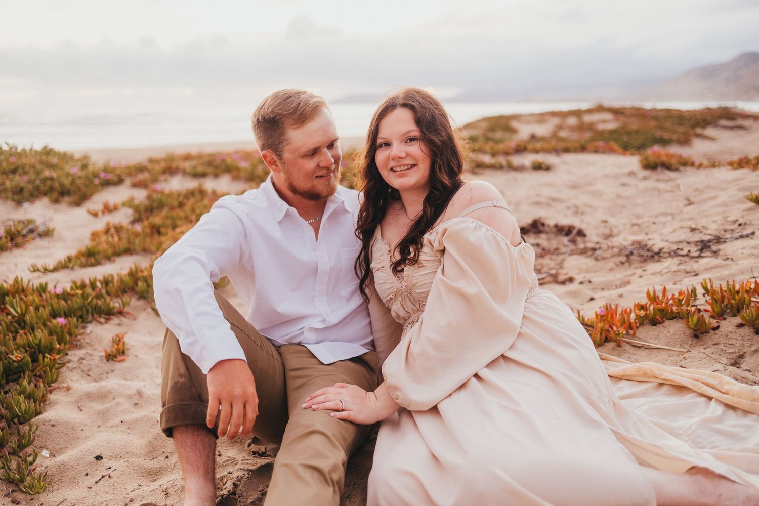 Couple in matching white and beige outfits share tender moment on coastal rocks at golden hour.