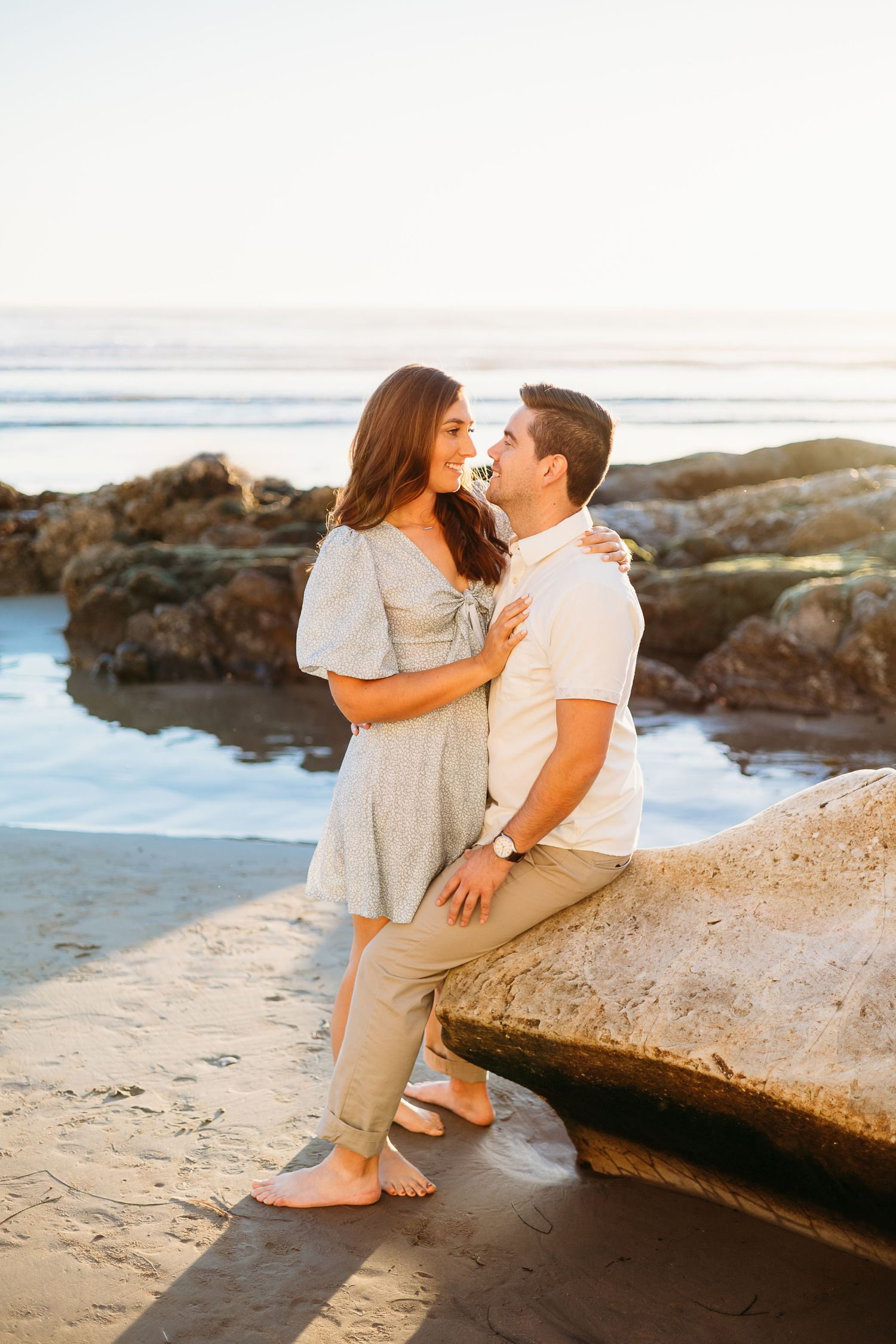 A couple embraces on a rocky beach with ocean waves and sunset in the background.
