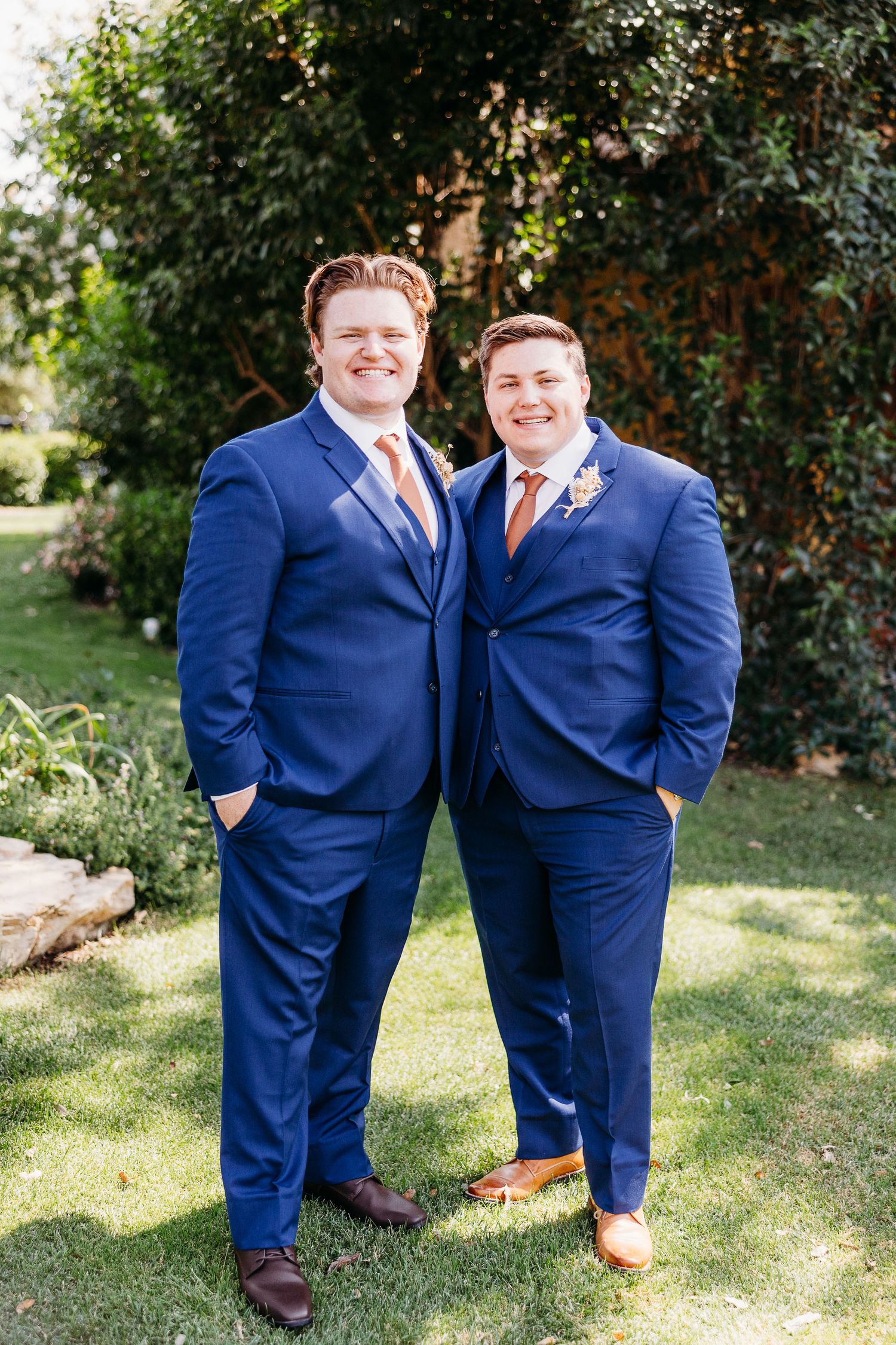 Two groomsmen in matching navy suits with orange ties pose together in a garden setting.