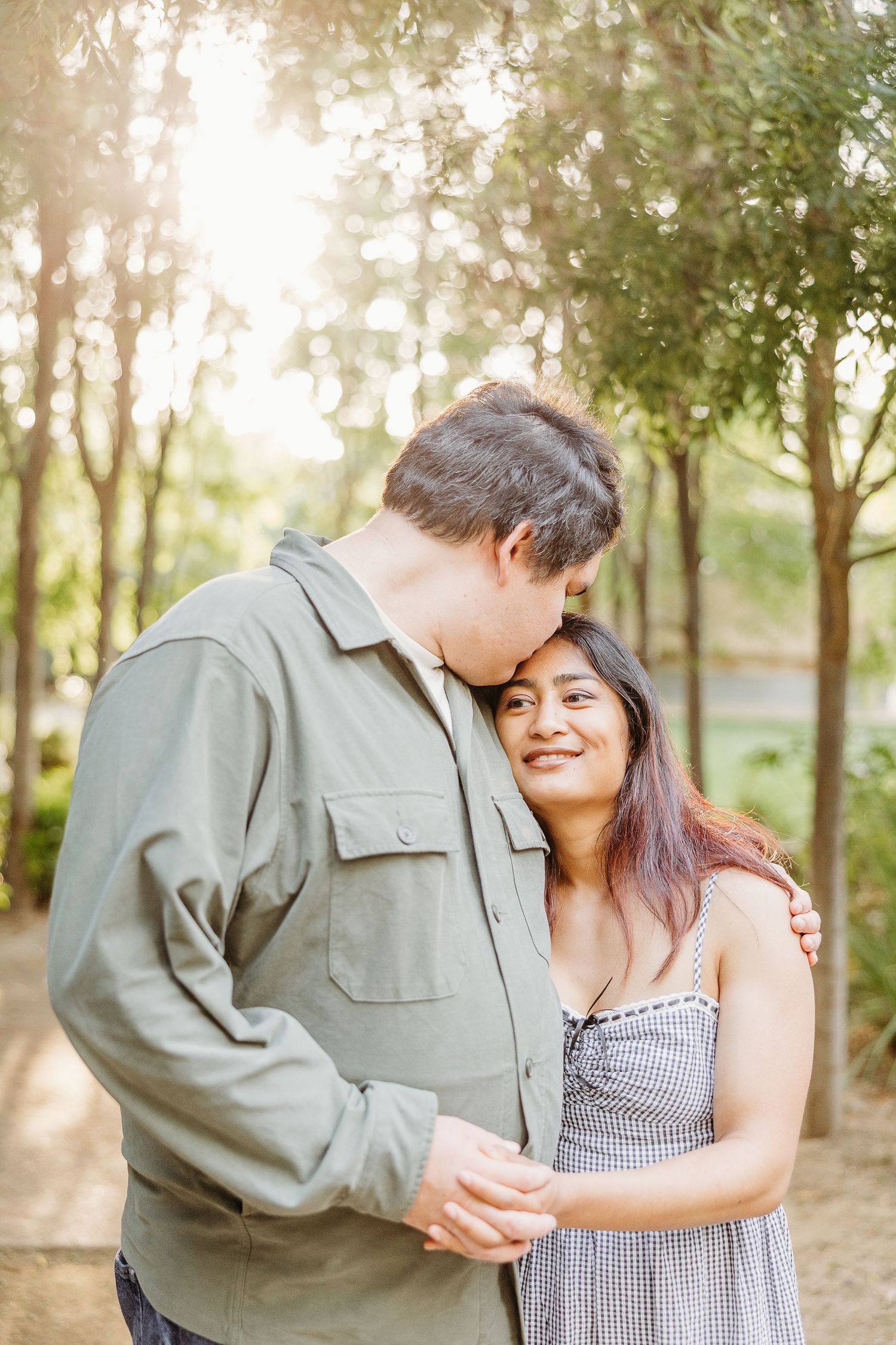 A couple embraces lovingly in a sunlit forest setting wearing casual attire.