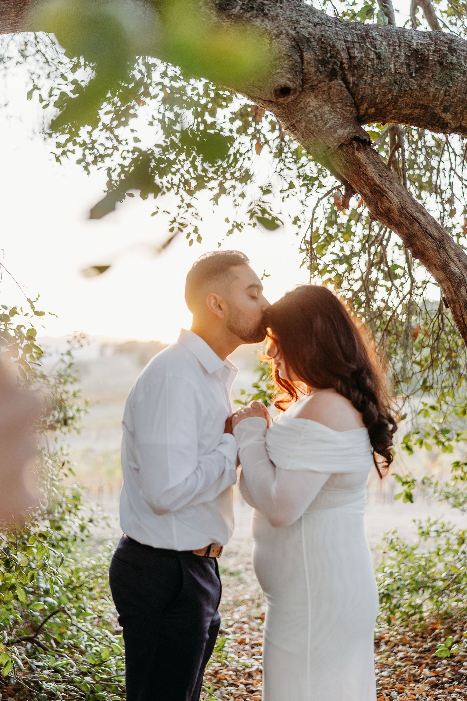 Couple sharing romantic moment under tree branches with sunlight filtering through leaves.