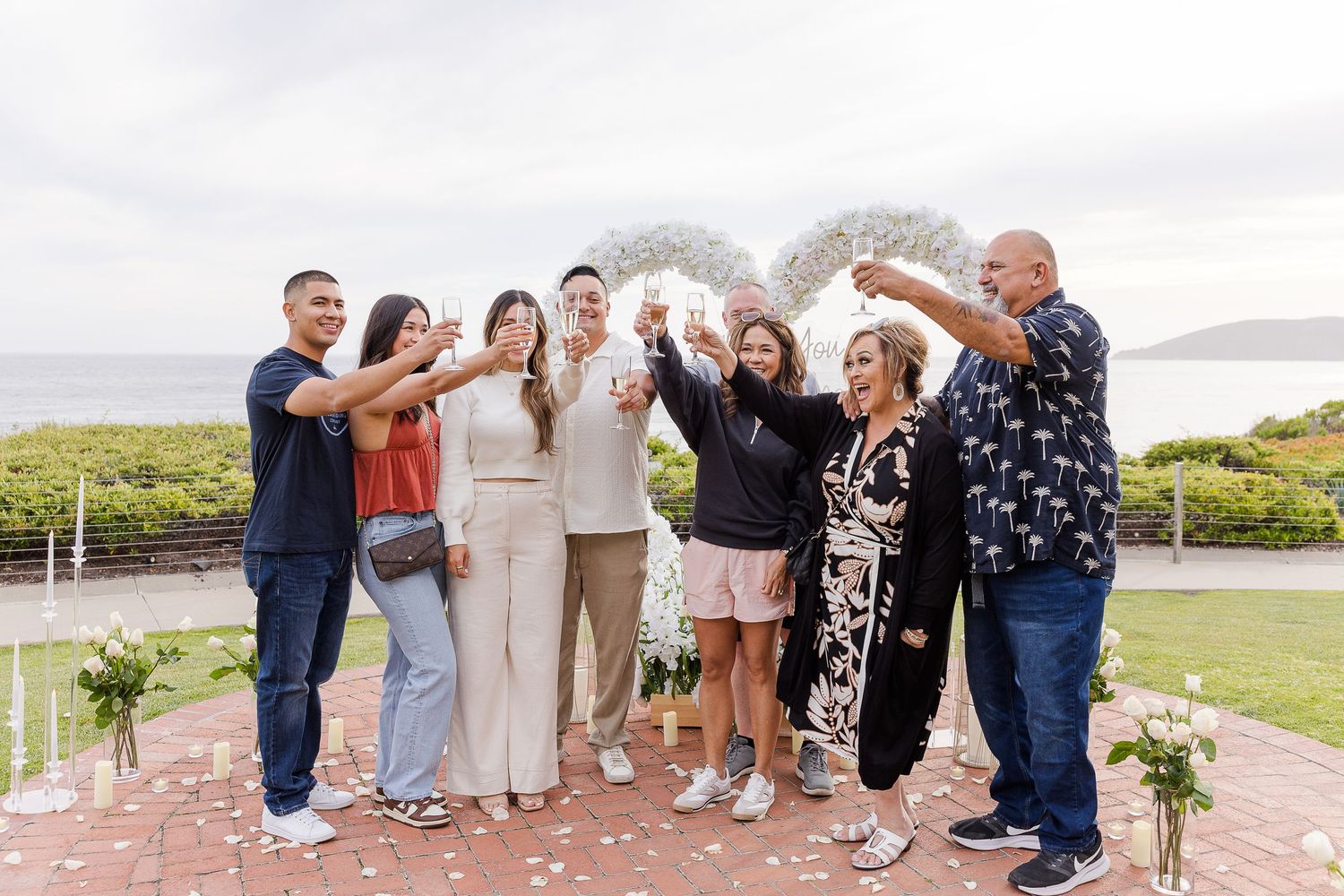 Group photo of wedding party and guests posing together outdoors with scenic ocean view in background.