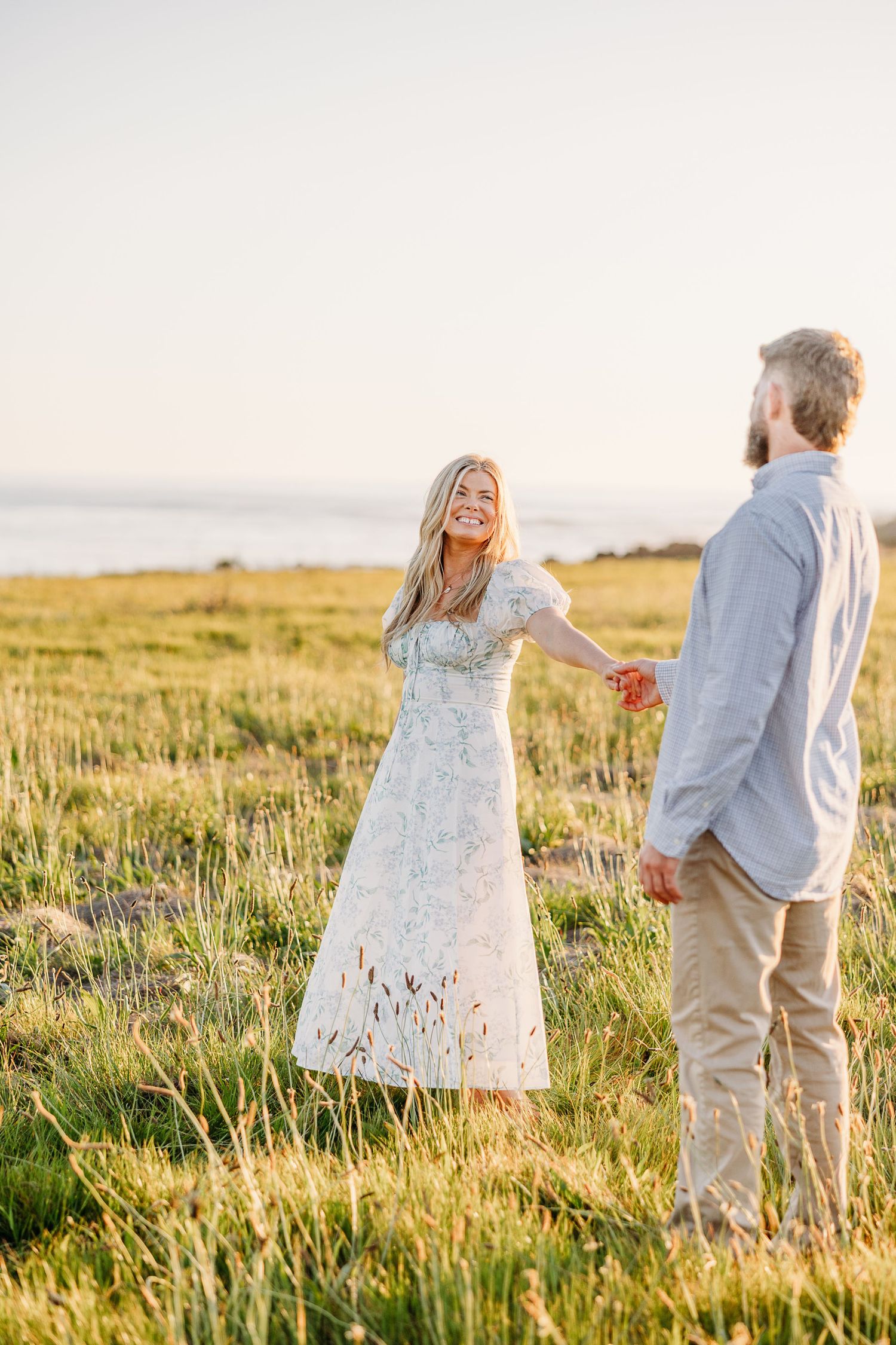 Couple walks through a sun-drenched meadow of tall grass during golden hour in romantic countryside setting.
