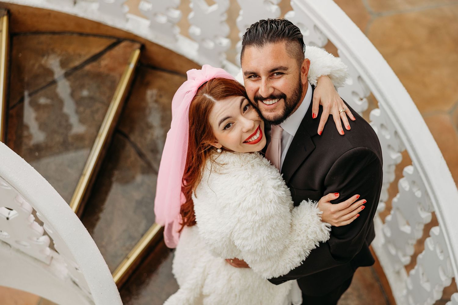 A happy stylish couple embraces on a staircase, with the person wearing a white furry jacket and pink hair.