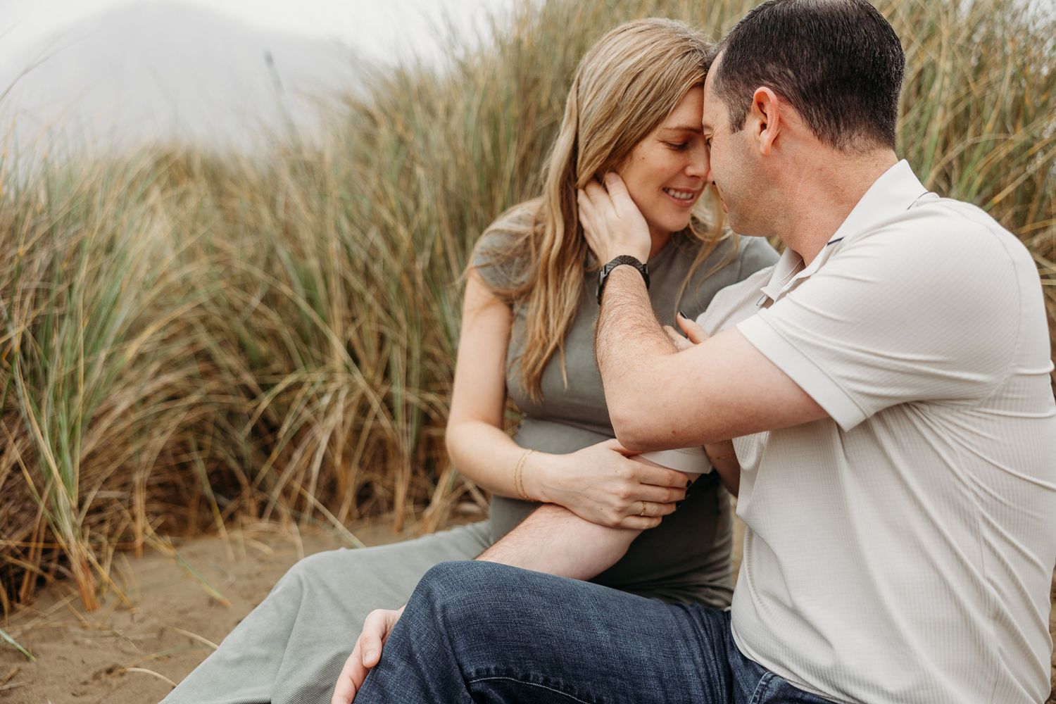 Romantic maternity photo of couple sitting in beach grass during golden hour.