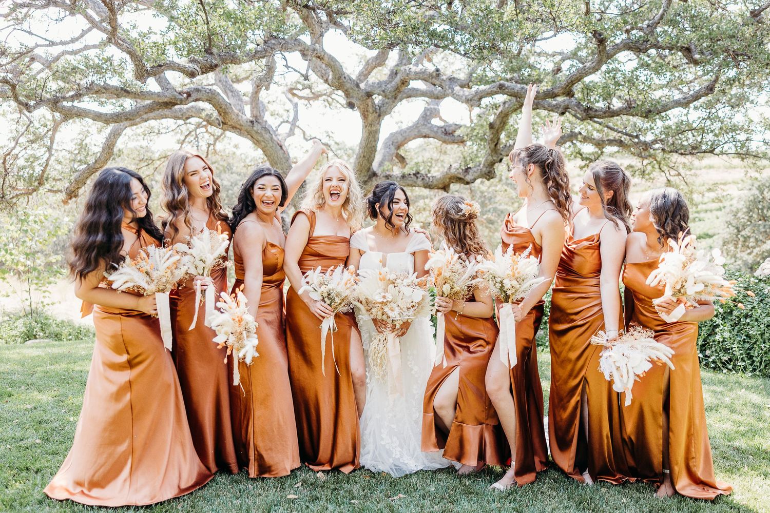 Bridesmaids in flowing orange dresses gather with bride for outdoor wedding photo.