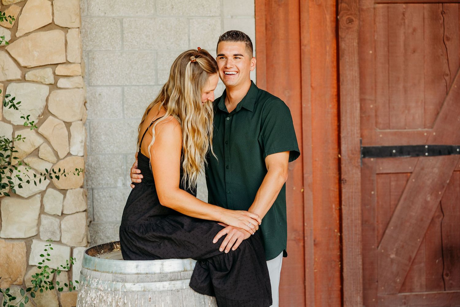 Couple embracing near a rustic barn door with stone wall and natural lighting.