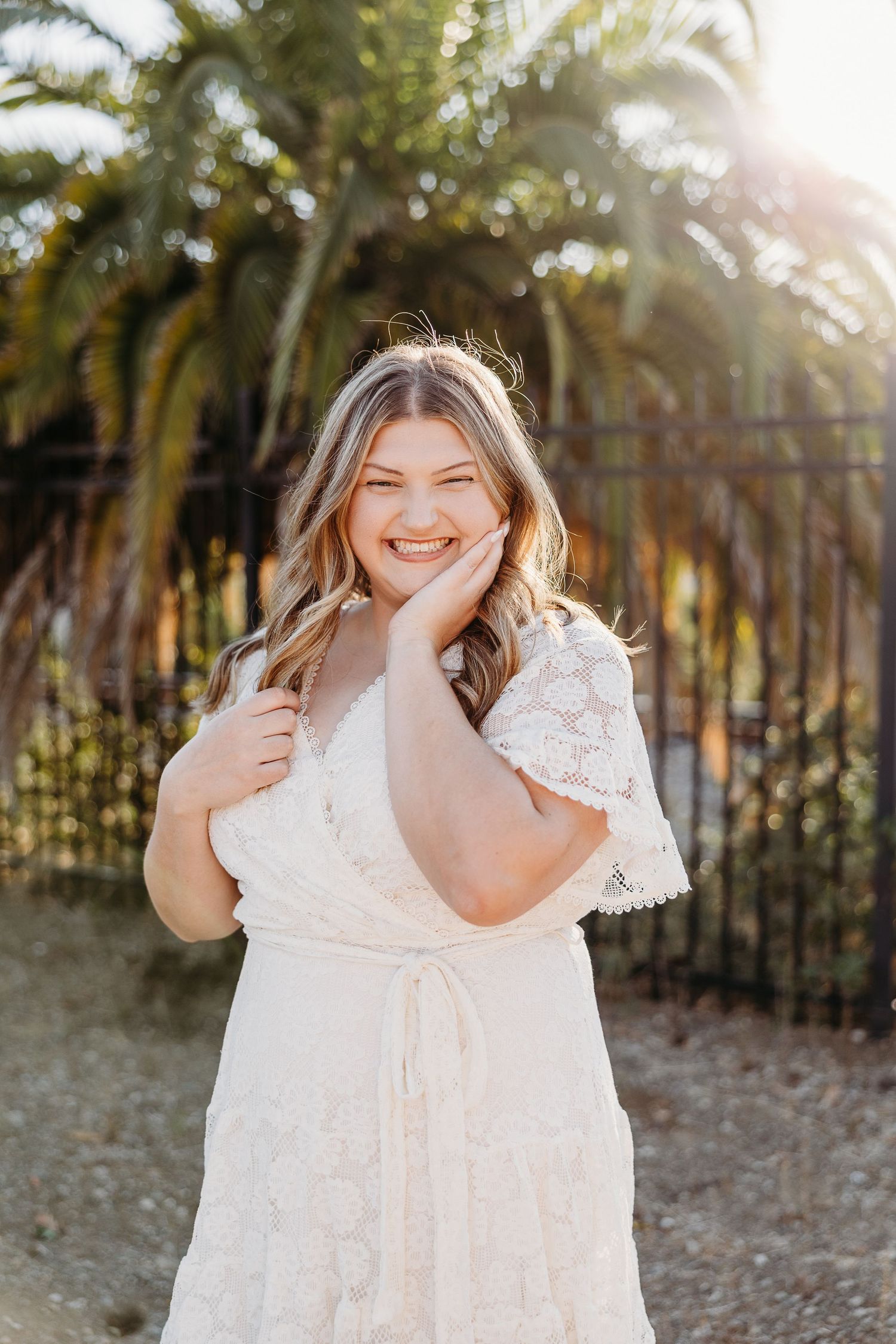 Someone in a white lace dress poses joyfully against a backdrop of palm trees at sunset.