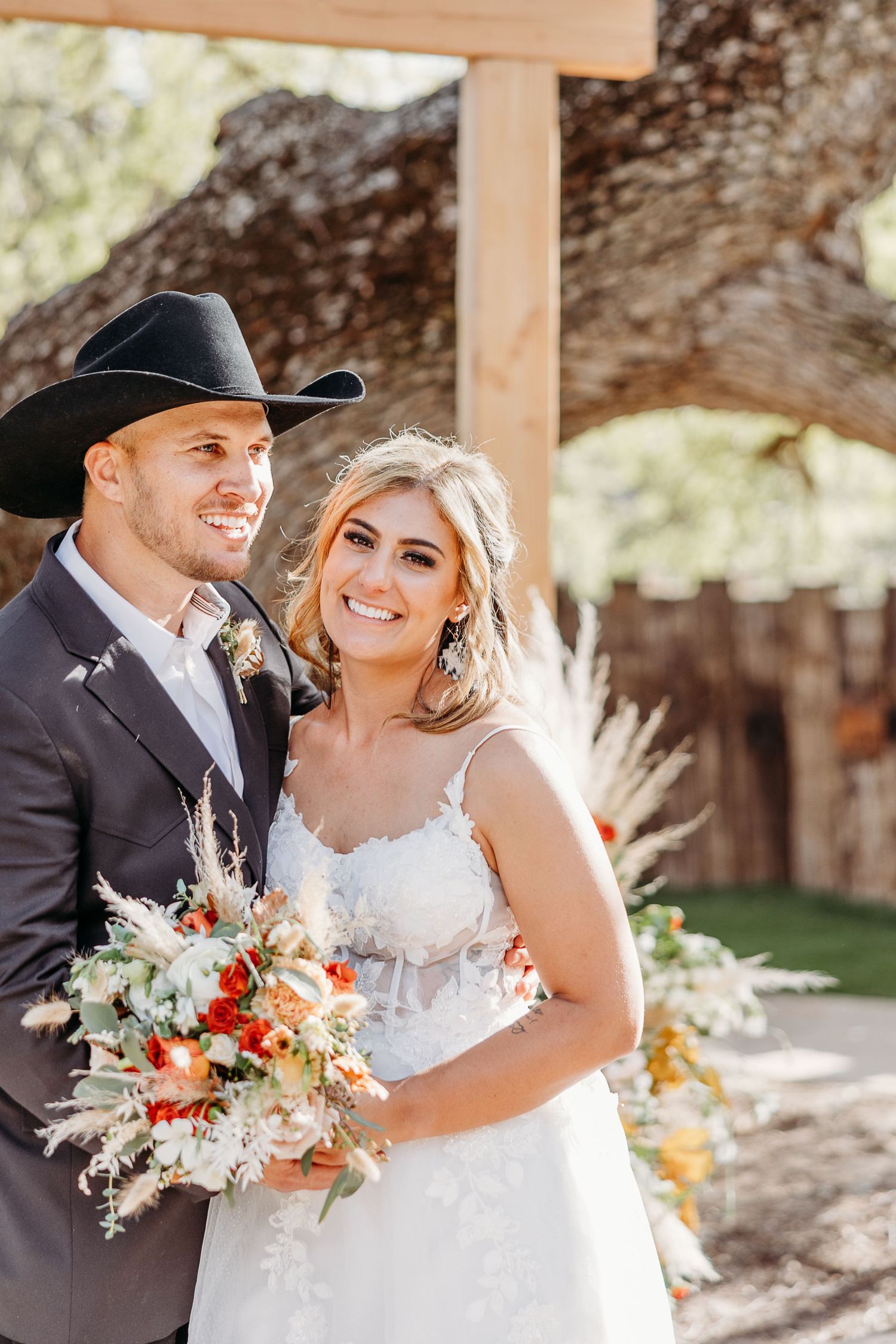 A bride in white lace dress holds vibrant fall bouquet next to groom wearing black cowboy hat under wooden pergola.