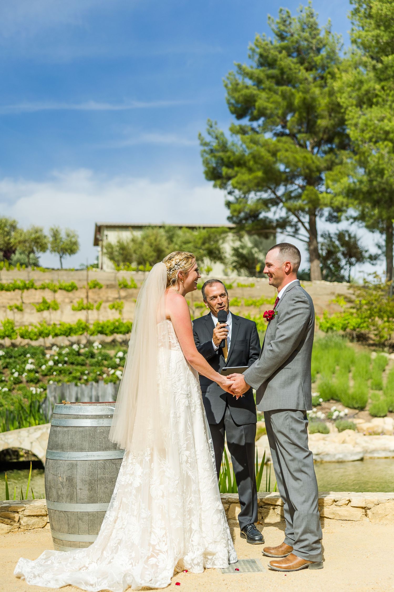 Outdoor wedding ceremony with wine barrels and scenic landscaping on a sunny day.