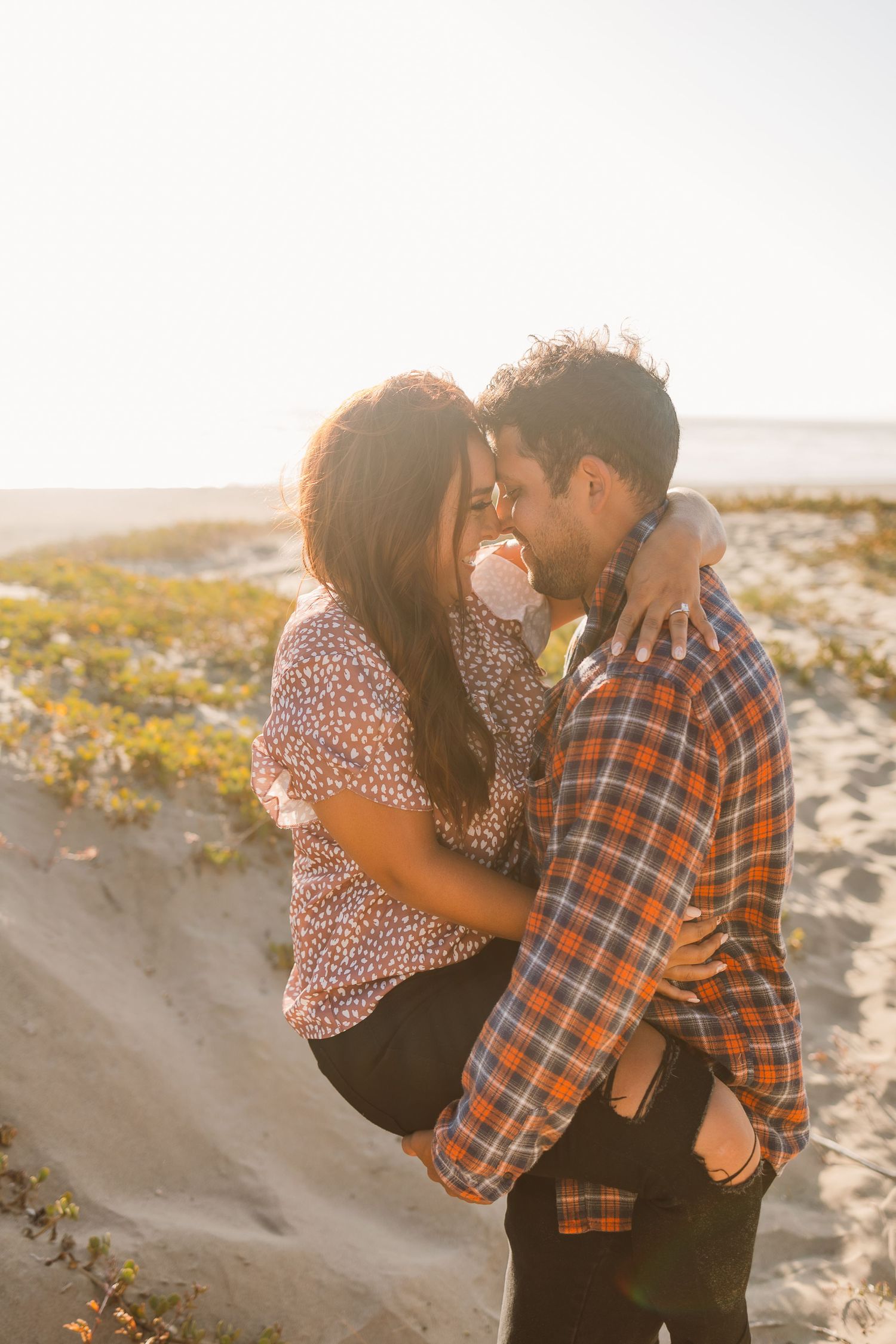 Couple sharing tender moment on sandy beach with vegetation in background.