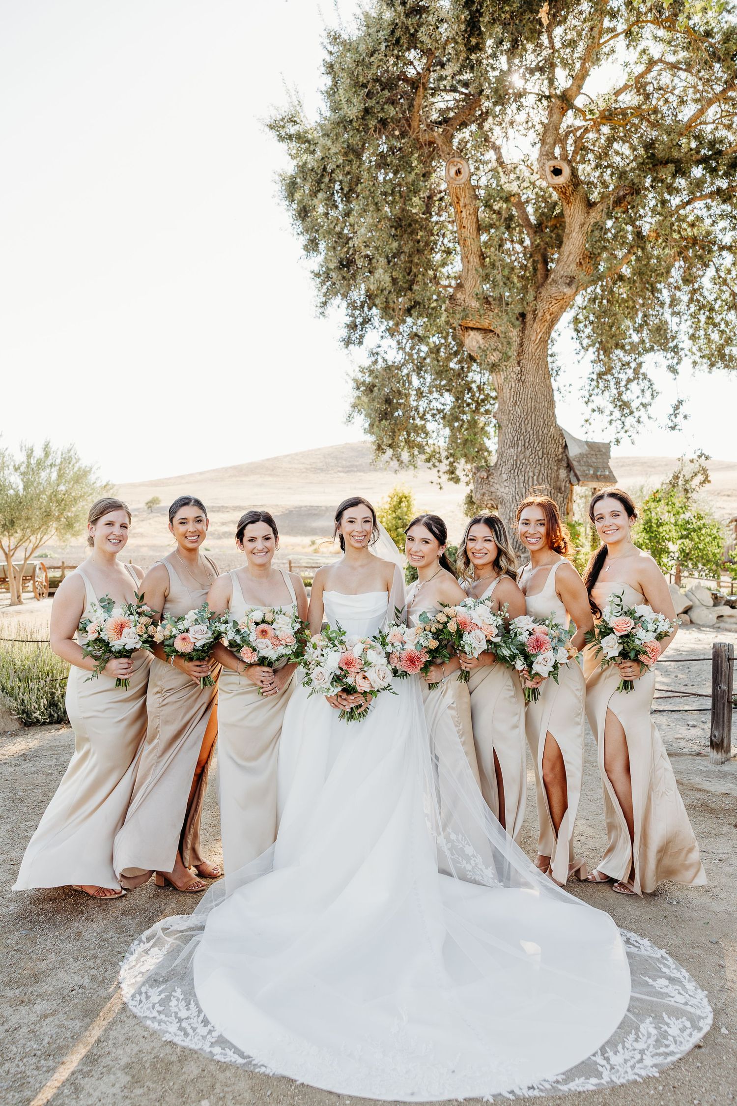 Bridesmaids in champagne dresses pose outdoors with floral bouquets featuring blush and white flowers.
