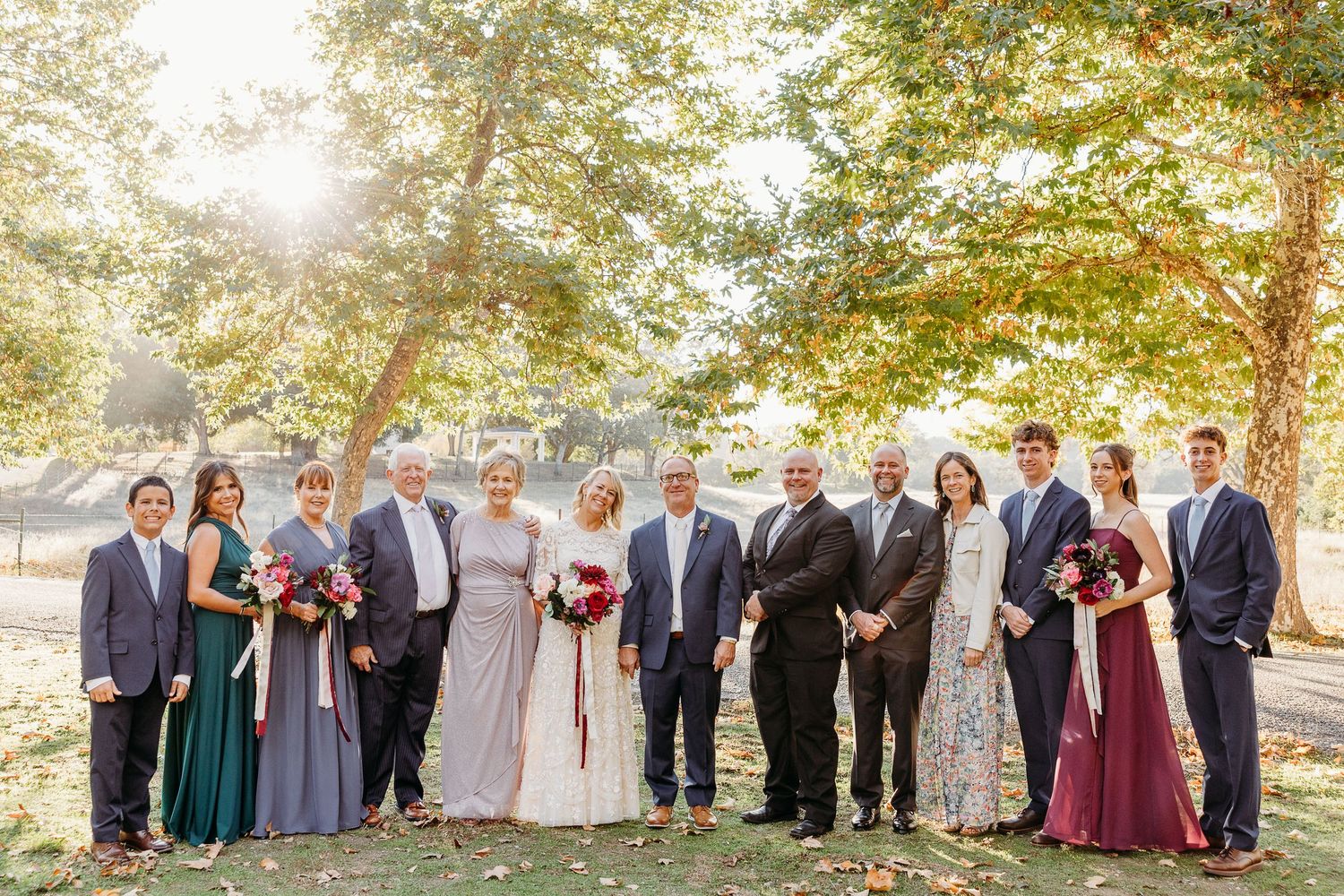 Wedding party poses for group photo in autumn outdoor setting with golden sunlight.